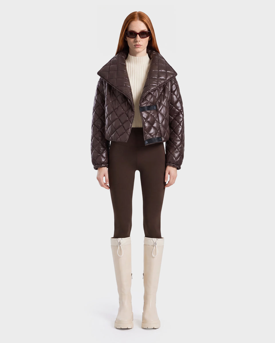 A woman stands facing forward against a plain light background, embodying active versatility in the Rudsak KALEE - 8124005 Espresso jacket styled with a cream turtleneck, brown pants, cream knee-high boots, and sunglasses for a chic and functional look.