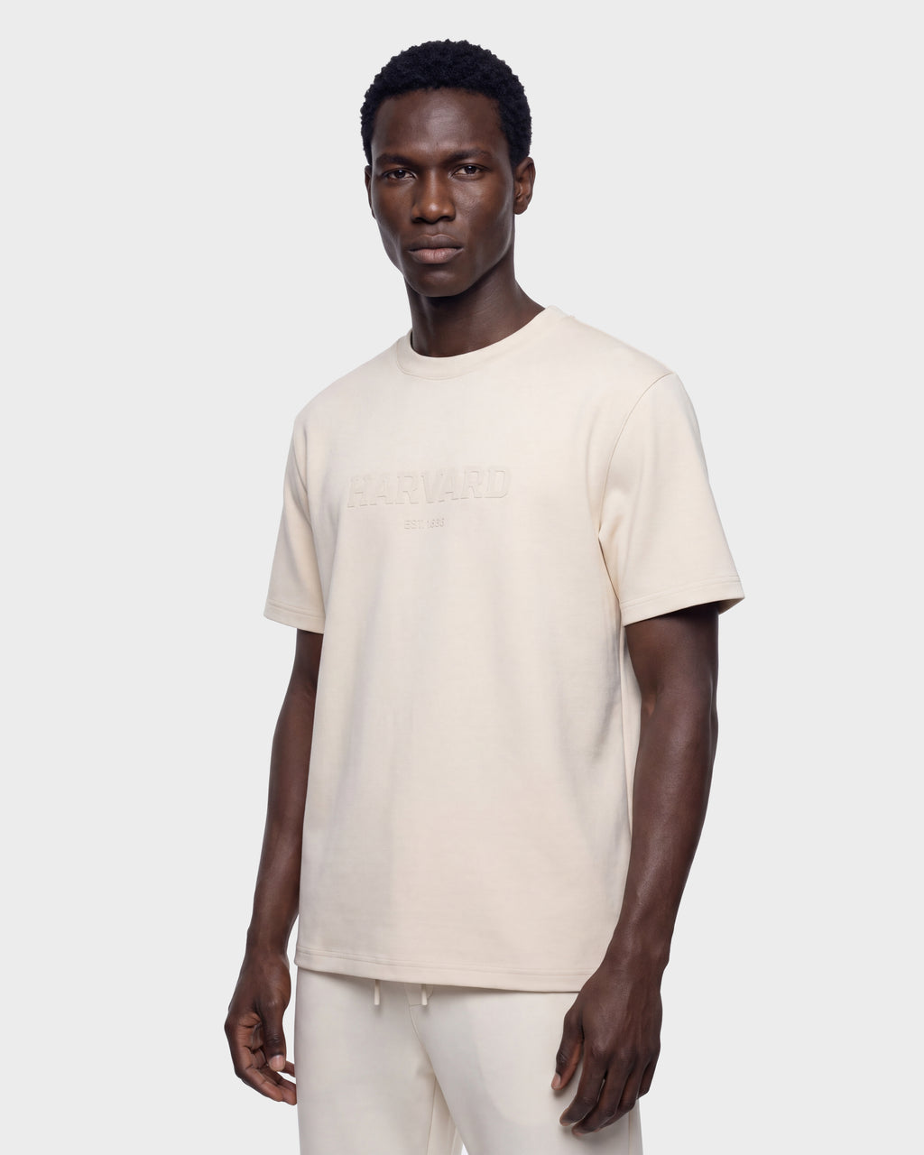 A man wears the Rudsak KARTER R HV - H525507 Vanilla limited edition T-shirt, featuring HARVARD EST. 1636 embossed on the front, paired with matching beige pants and standing against a plain light background—an effortlessly cool take on modern streetwear.