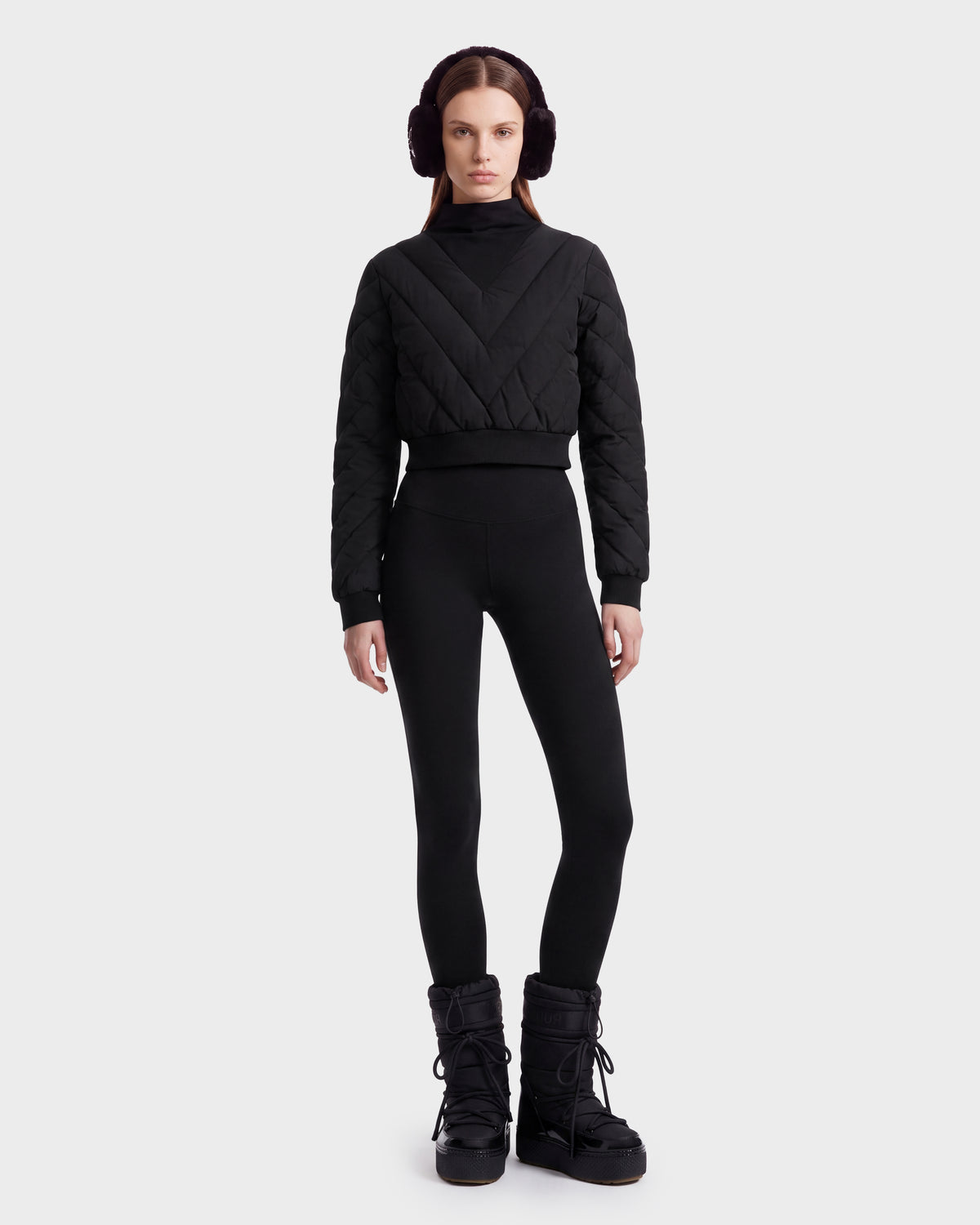 A person stands against a plain background wearing black earmuffs, sleek black leggings, and winter boots, styled with the Rudsak KATYA - 8526531 Black quilted sweater—perfect for an après-ski look.