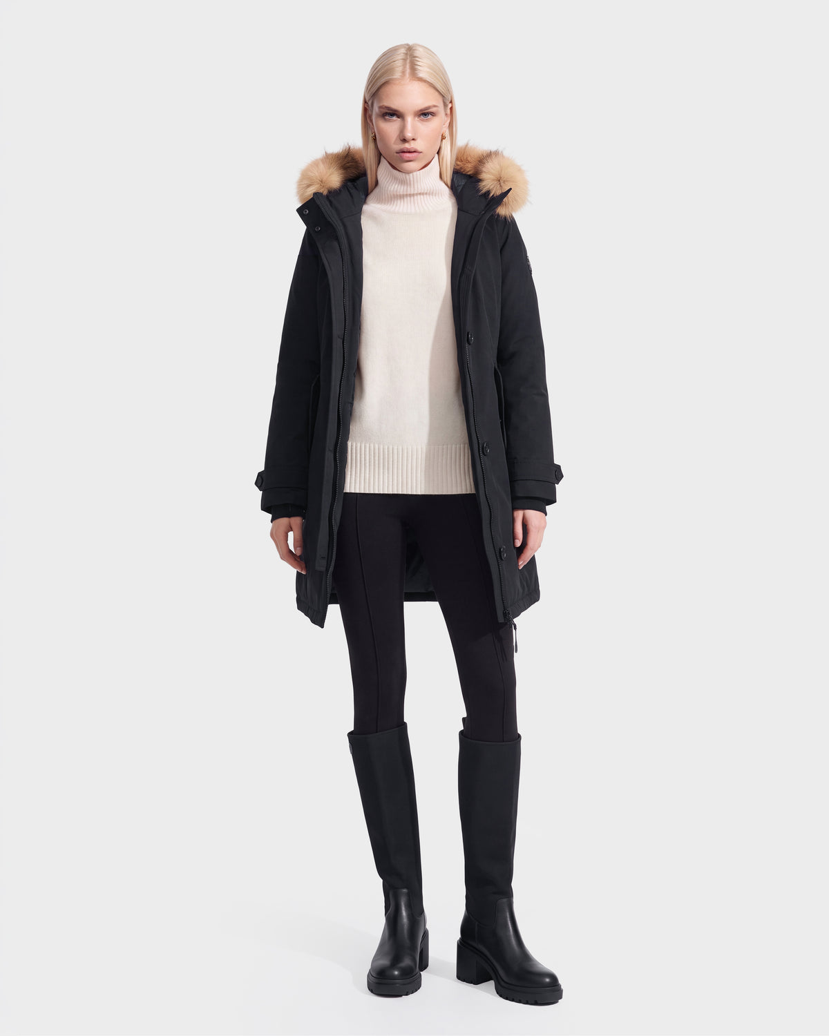 A person stands facing forward against a plain light background, wearing the Rudsak KEYSHIA - 8126517 Black parka with a fur-lined hood, a cream turtleneck sweater, black leggings, and black knee-high boots.