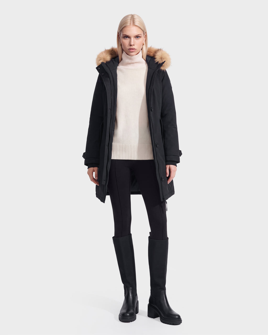 A person in the Rudsak KEYSHIA - 8126517 Black women’s down parka, a cream turtleneck sweater, black leggings, and knee-high boots stands facing forward against a plain light background.