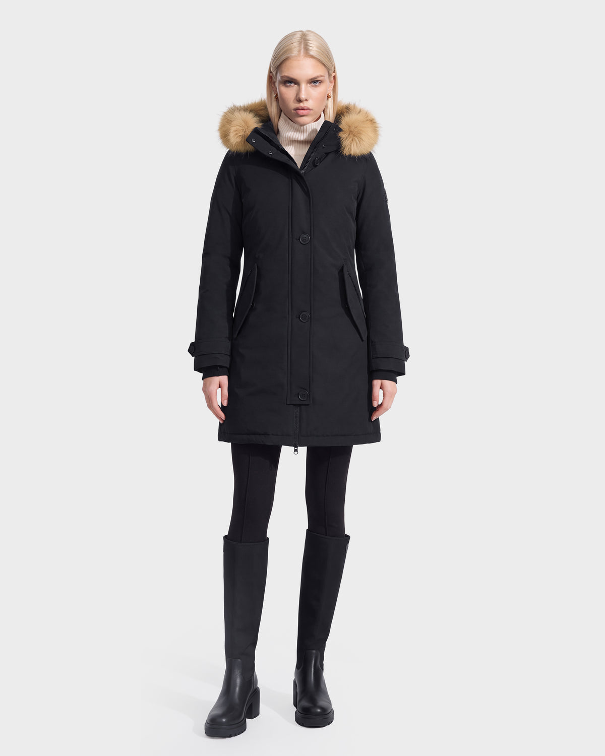 A woman wearing the Rudsak KEYSHIA - 8126517 Black parka with a fur-trimmed hood, paired with black leggings and black knee-high boots, stands against a plain white background.