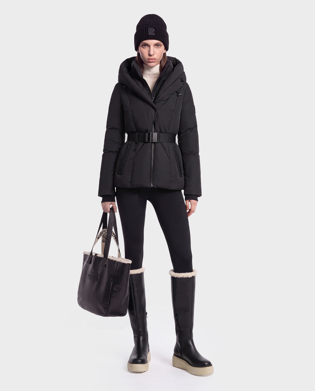 A woman wearing a black Rudsak KOLAD - 8124528 puffer jacket with RDS-certified down insulation, black pants, tall black boots with white soles, a black beanie, and a white turtleneck is holding a black tote bag with vegan leather trims while standing against a plain white background.