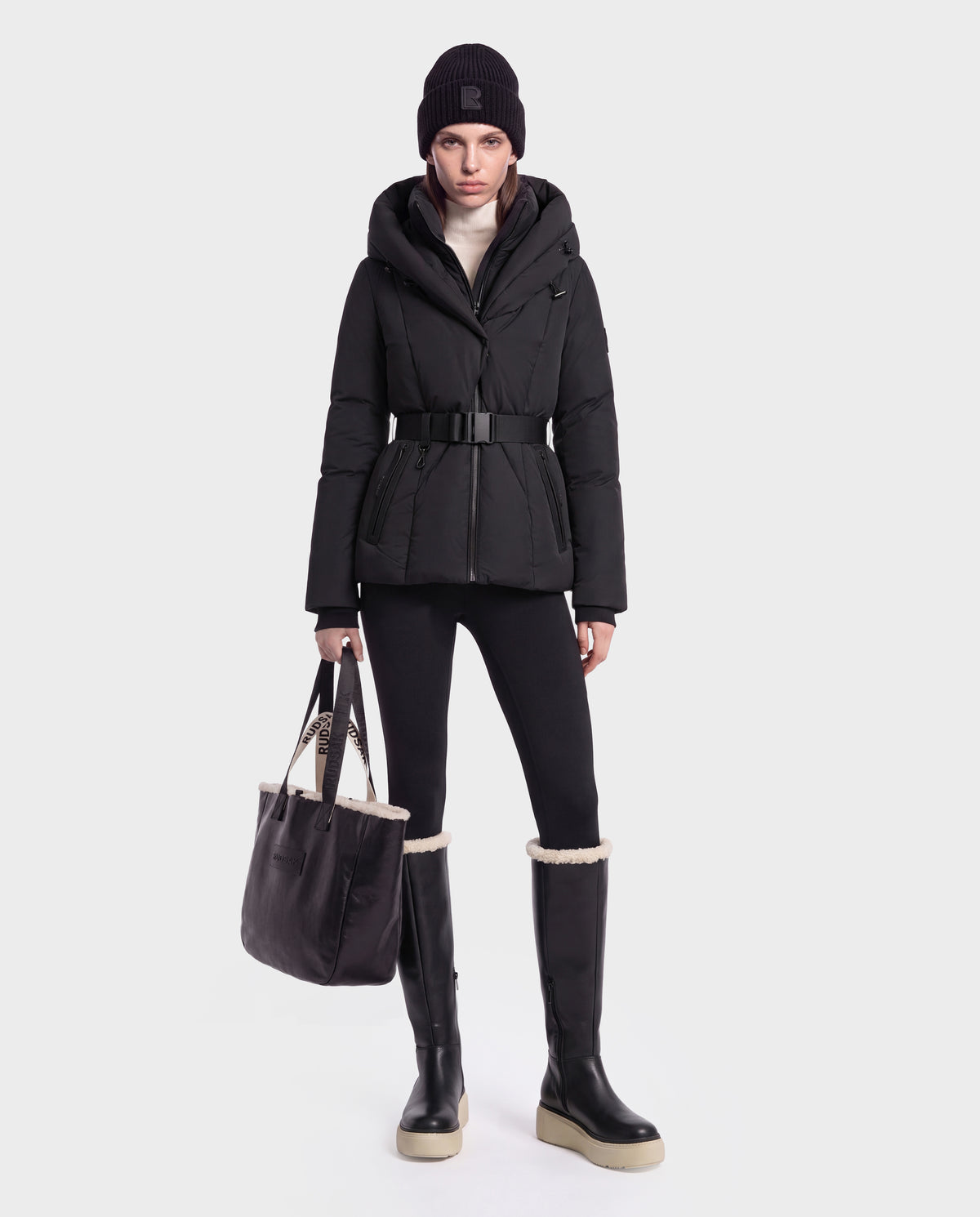 A woman wearing a black Rudsak KOLAD - 8124528 puffer jacket with RDS-certified down insulation, black pants, tall black boots with white soles, a black beanie, and a white turtleneck is holding a black tote bag with vegan leather trims while standing against a plain white background.