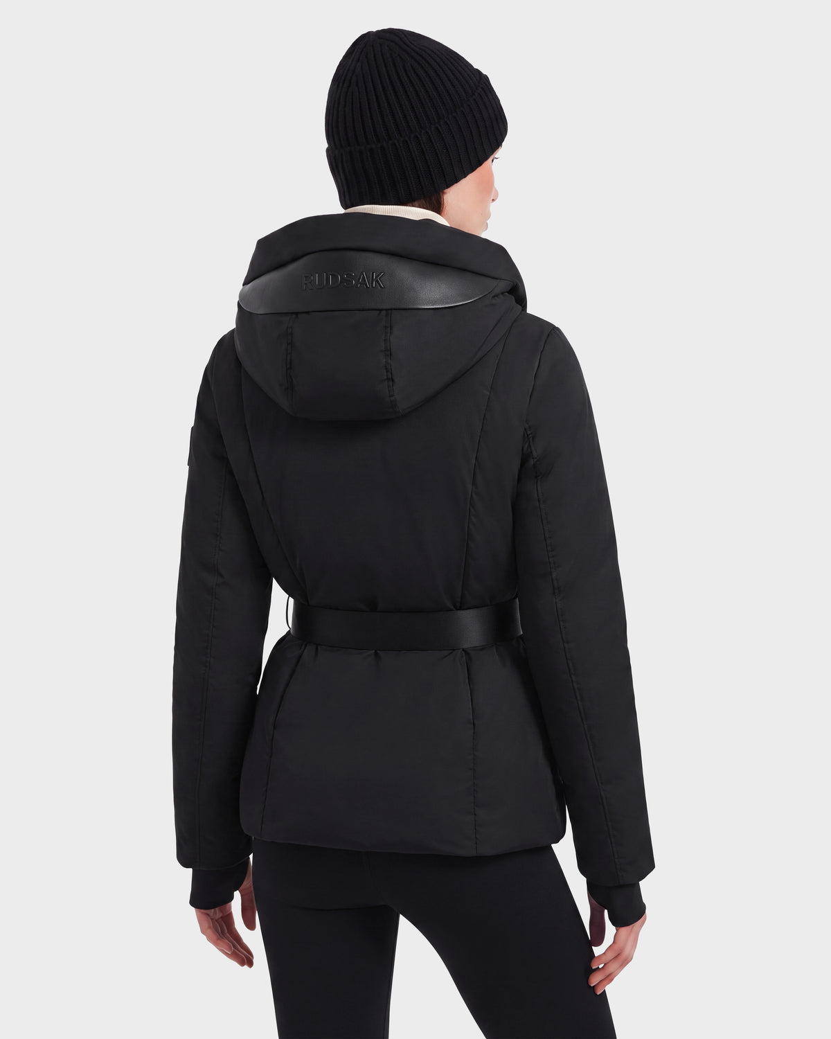 A person wearing the Rudsak KOLAD - 8124528 Black hooded jacket with vegan leather trims and a belt, paired with a black knit beanie, is shown from the back against a plain light background.