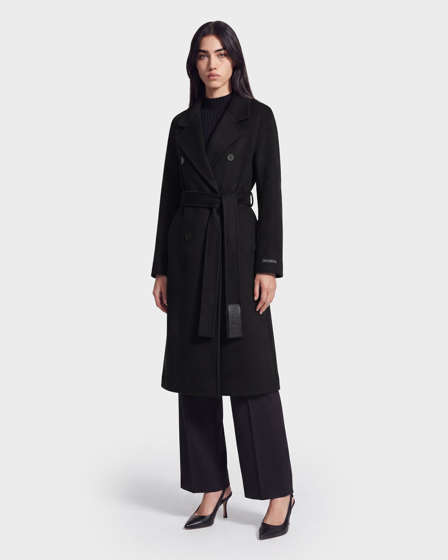A woman stands facing forward in the KOURT W - 8124702 Black coat by Rudsak, a luxurious long black double-breasted piece crafted from a premium wool and cashmere blend. She pairs it with black pants, black heels, and a black top against a plain light background.