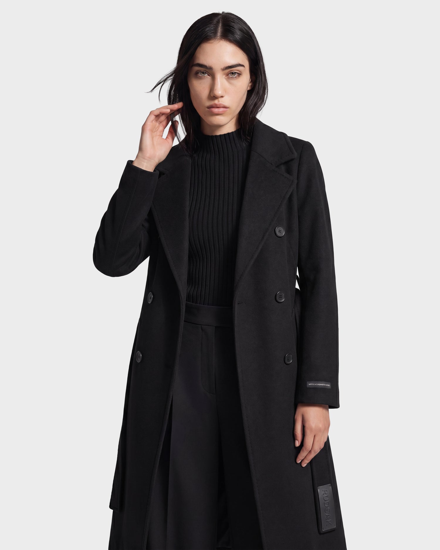 A woman with long dark hair wears the Rudsak KOURT W - 8124702 Black double-breasted coat, crafted from a luxurious wool and cashmere blend, layered over a black ribbed sweater. She poses with one hand near her face against a plain light background.