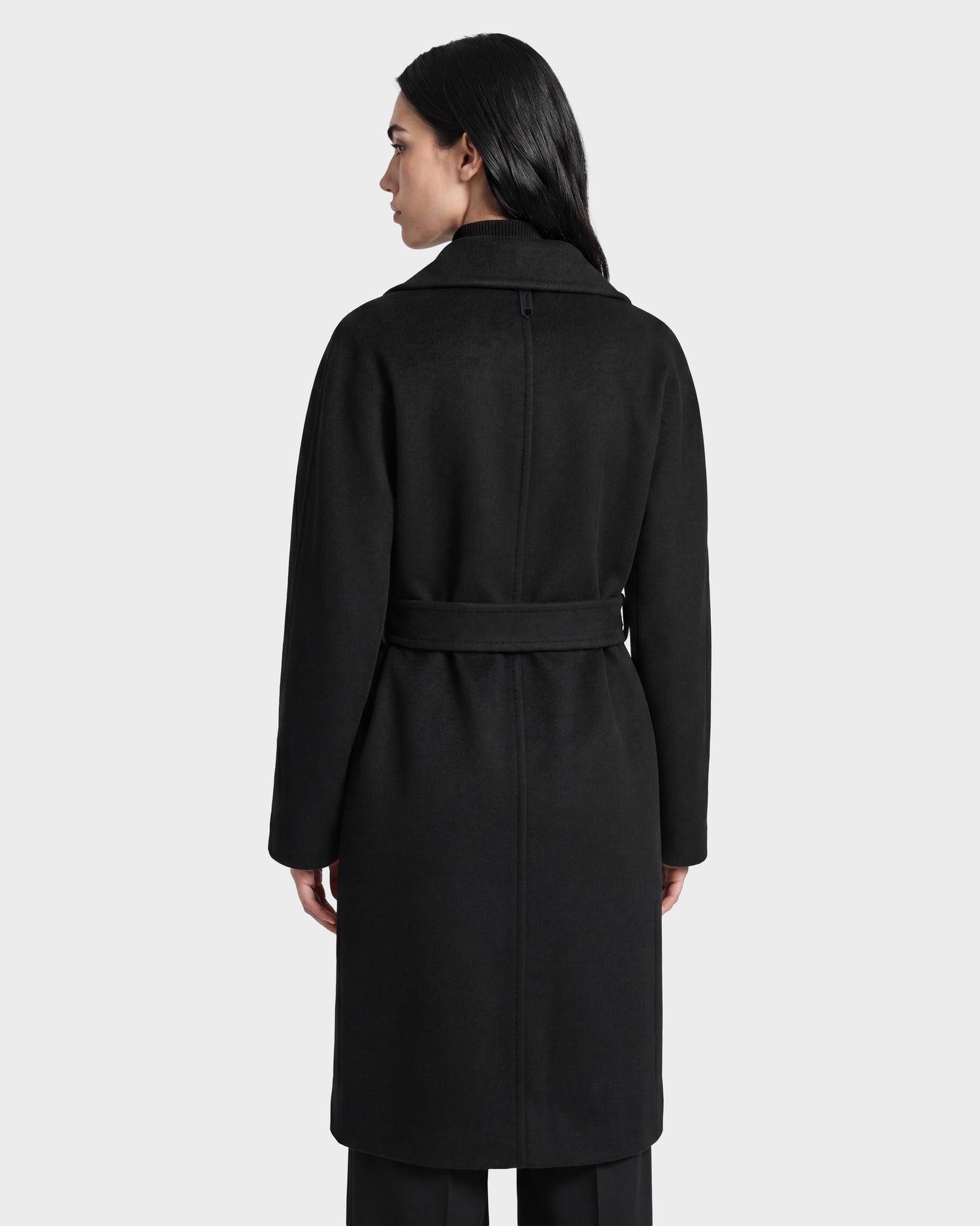 A woman with long dark hair is shown from the back, wearing the Rudsak KOURT W - 8124702 Black belted wool coat and black pants, standing against a plain light gray background.