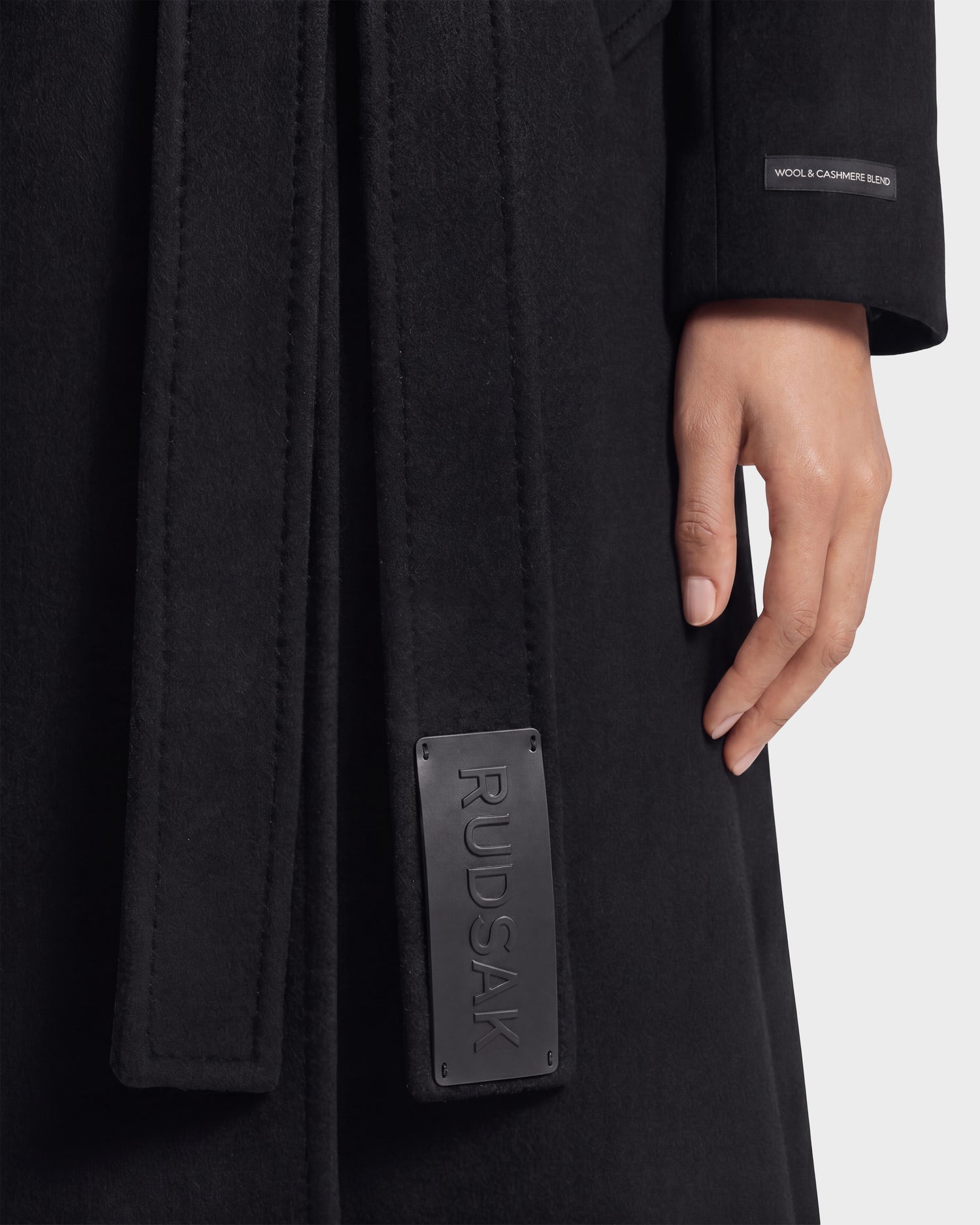 A close-up of a person wearing the Rudsak KOURT W - 8124702 Black women's belted wool coat. The belt is accented with a leather patch labeled “RUDSAK,” while a small tag on the sleeve reads “WOOL & CASHMERE BLEND.” Only one hand is visible, highlighting the luxurious texture and refined details of this double-breasted coat.