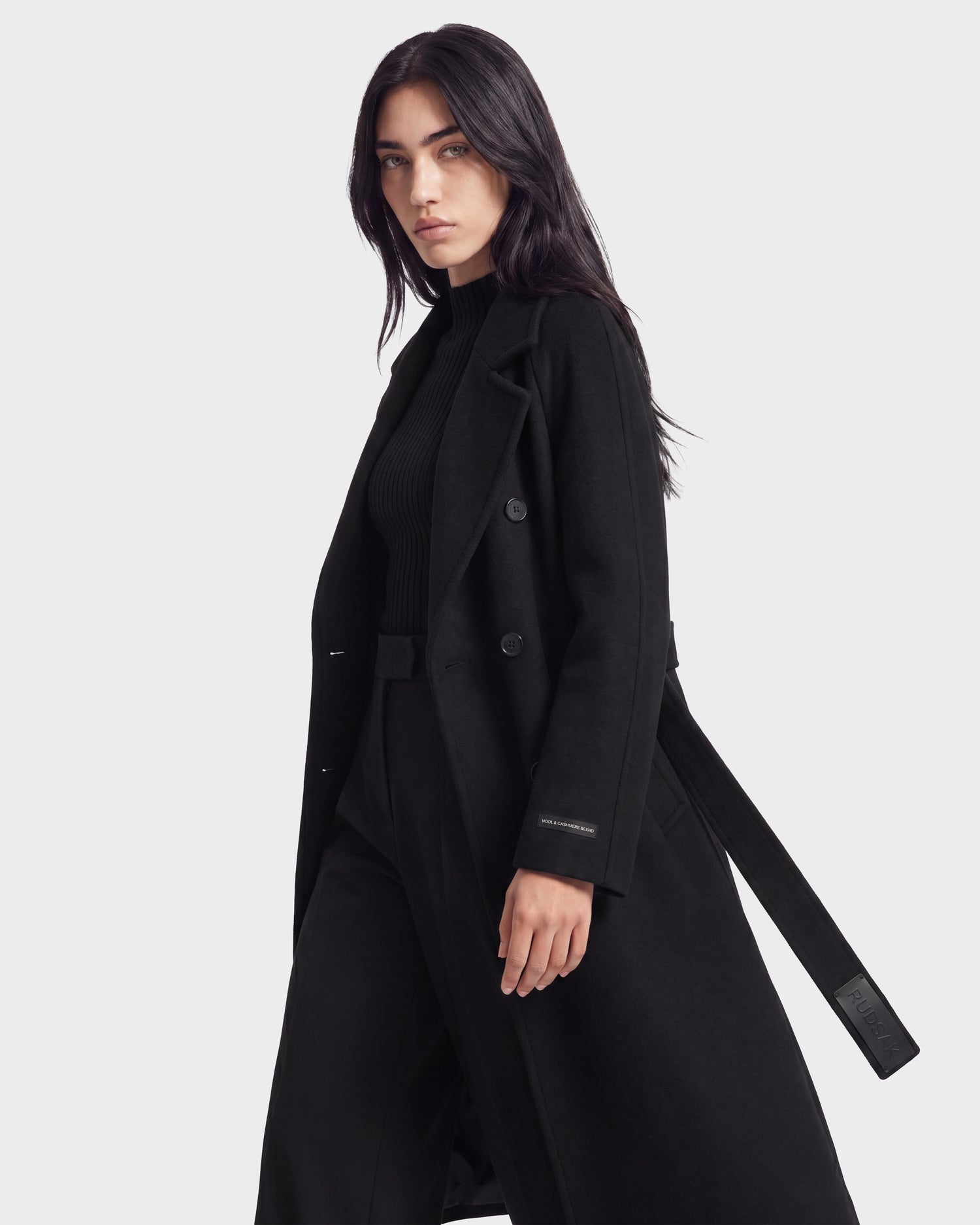 A woman with long dark hair wears the KOURT W - 8124702 Black women's belted wool coat by Rudsak over a black turtleneck and pants, standing against a plain white background and looking confidently at the camera. The luxurious wool and cashmere blend of the Rudsak coat adds an elegant touch to her sophisticated ensemble.