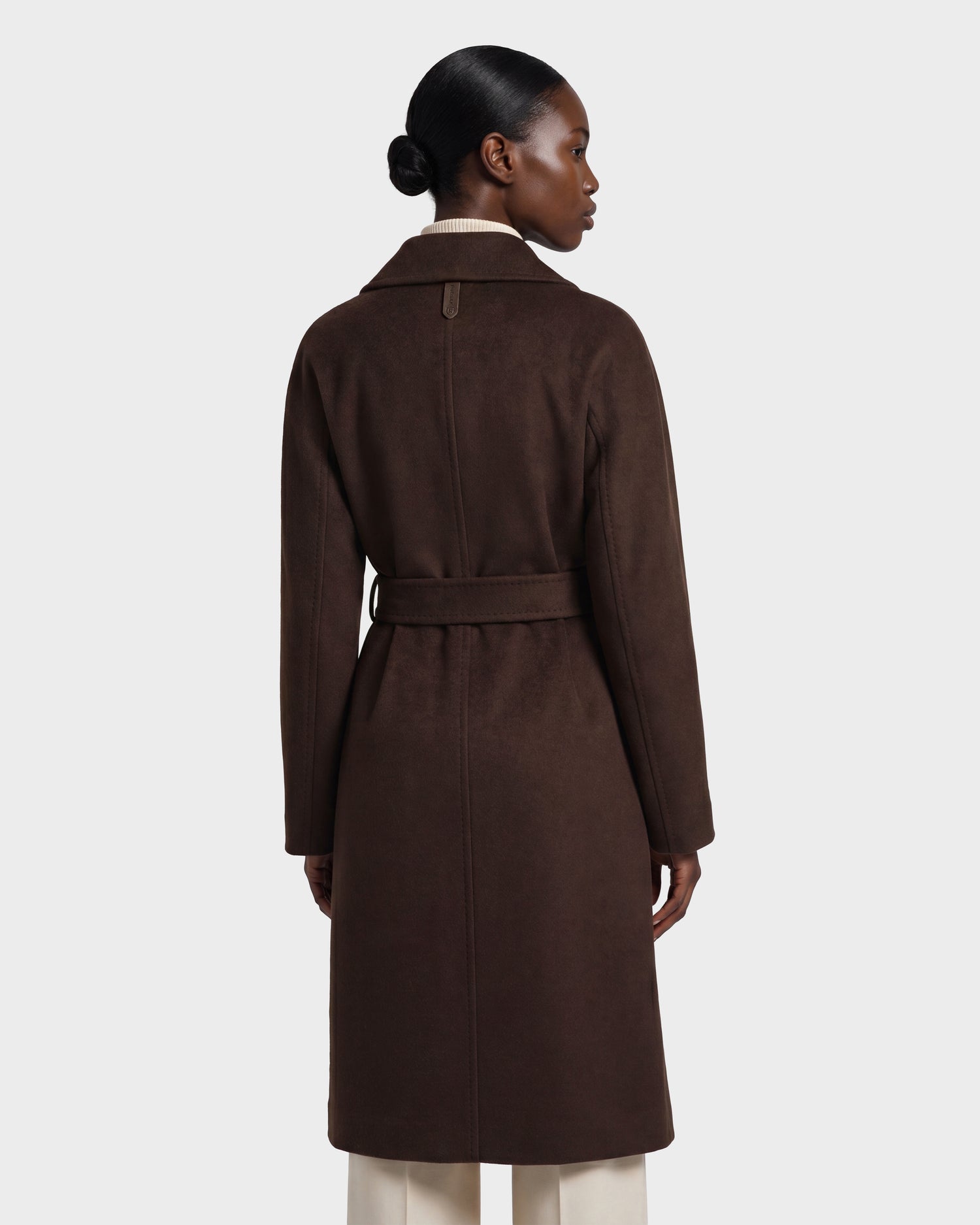 A person stands facing away, wearing the Rudsak KOURT W - 8124702 Espresso, a long dark brown belted wool coat with a structured collar, over light pants. Their hair is styled in a low bun and the background is plain and light.