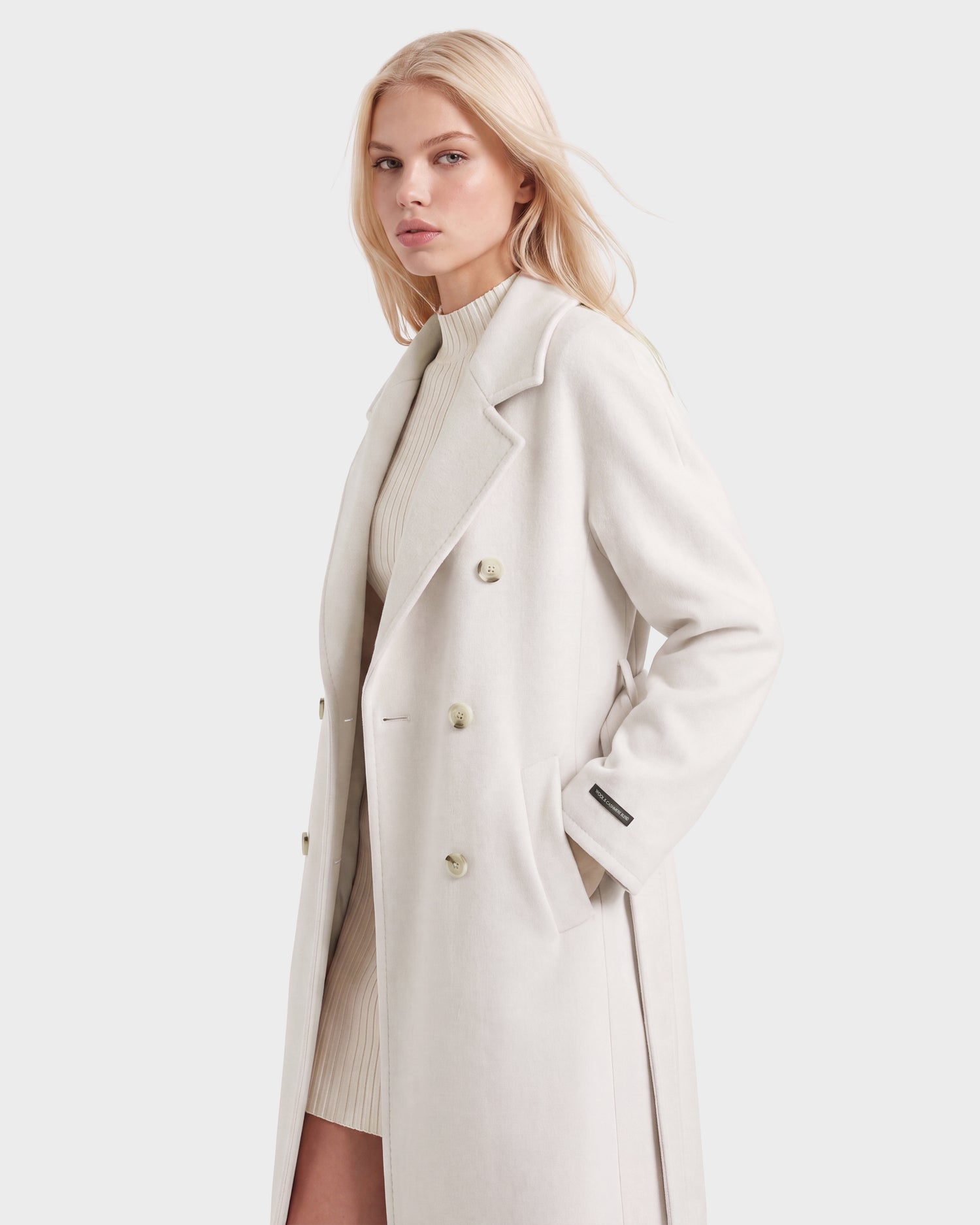 A woman with long blonde hair wears the Rudsak KOURT W - 8124702 Vanilla double-breasted coat over a ribbed turtleneck dress, standing against a plain light background.