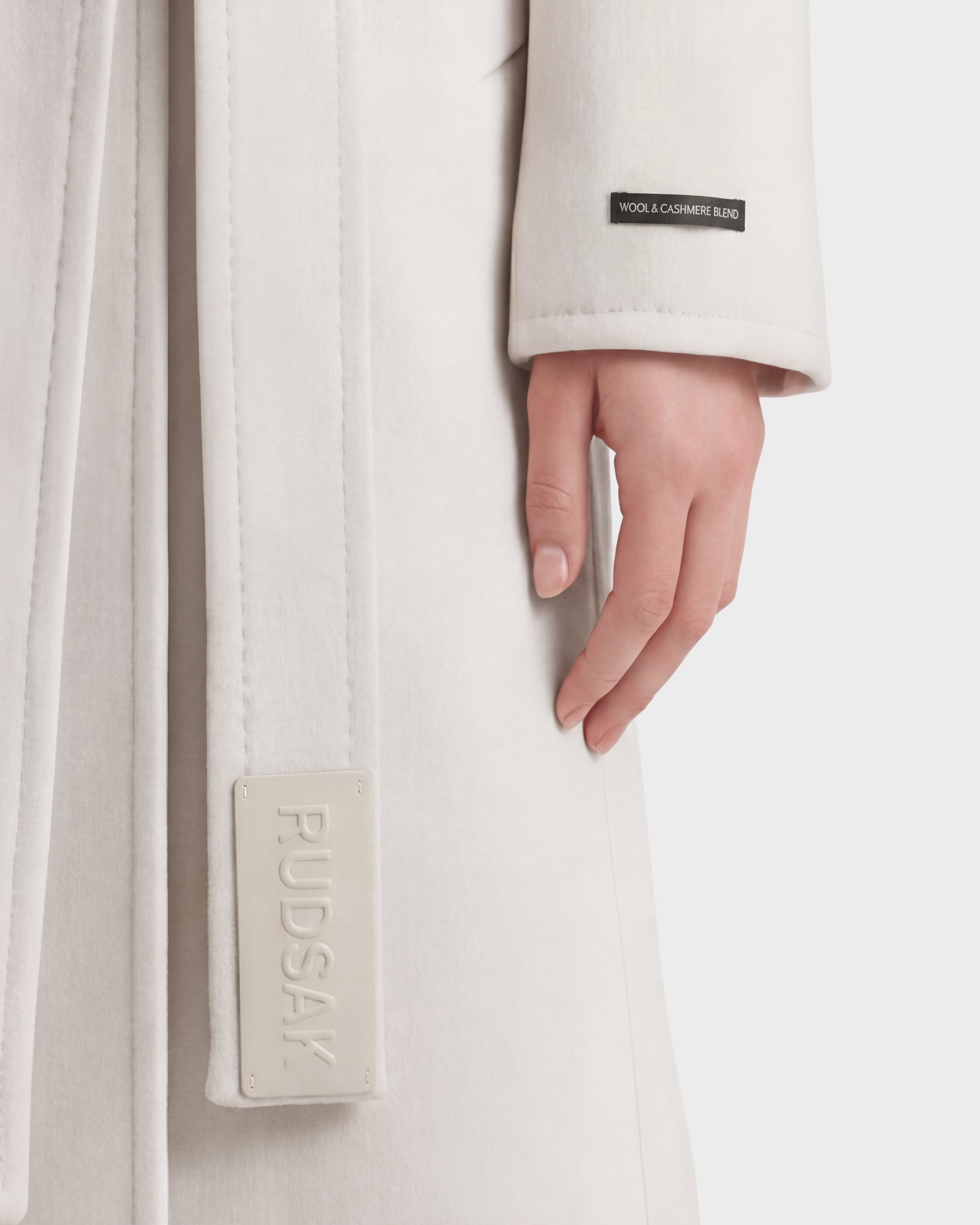 A close-up of a person wearing the KOURT W - 8124702 Vanilla women’s wool coat by Rudsak, featuring a belted design and showing the RUDSAK label along with a WOOL & CASHMERE BLEND tag near the sleeve; only their right hand and part of the light-colored coat are visible.