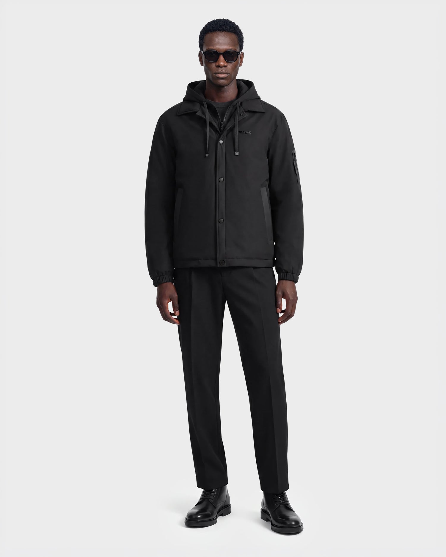 A man wearing sunglasses, the Rudsak LUCA - 6125009 black weather-resistant hooded men’s jacket, black pants, and black shoes stands against a plain white background.