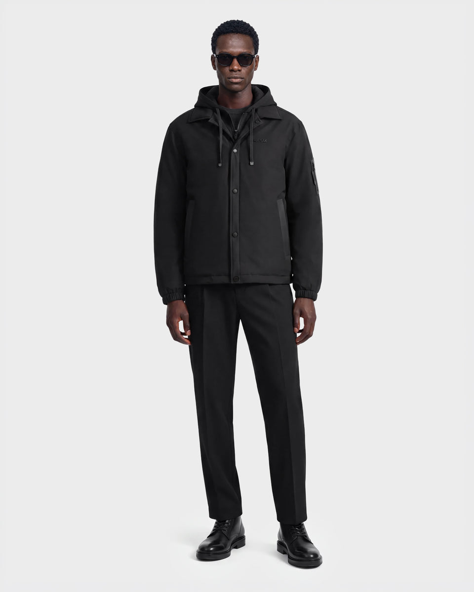 A man wearing sunglasses, the Rudsak LUCA - 6125009 black weather-resistant hooded men’s jacket, black pants, and black shoes stands against a plain white background.