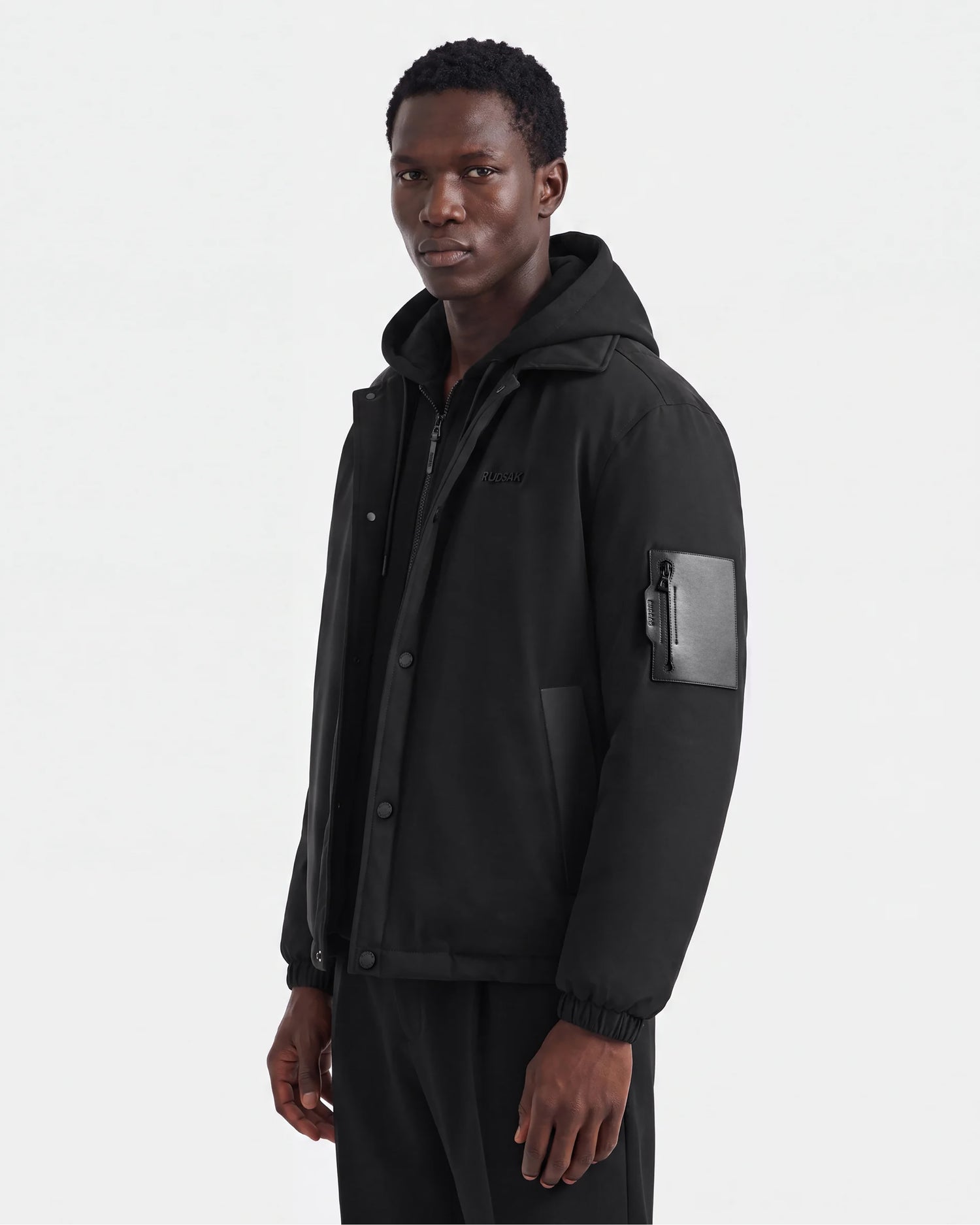 A man wearing the LUCA - 6125009 Black lightweight insulated men's jacket by Rudsak, featuring a zippered pocket on the left sleeve, stands facing slightly to the side against a plain white background.