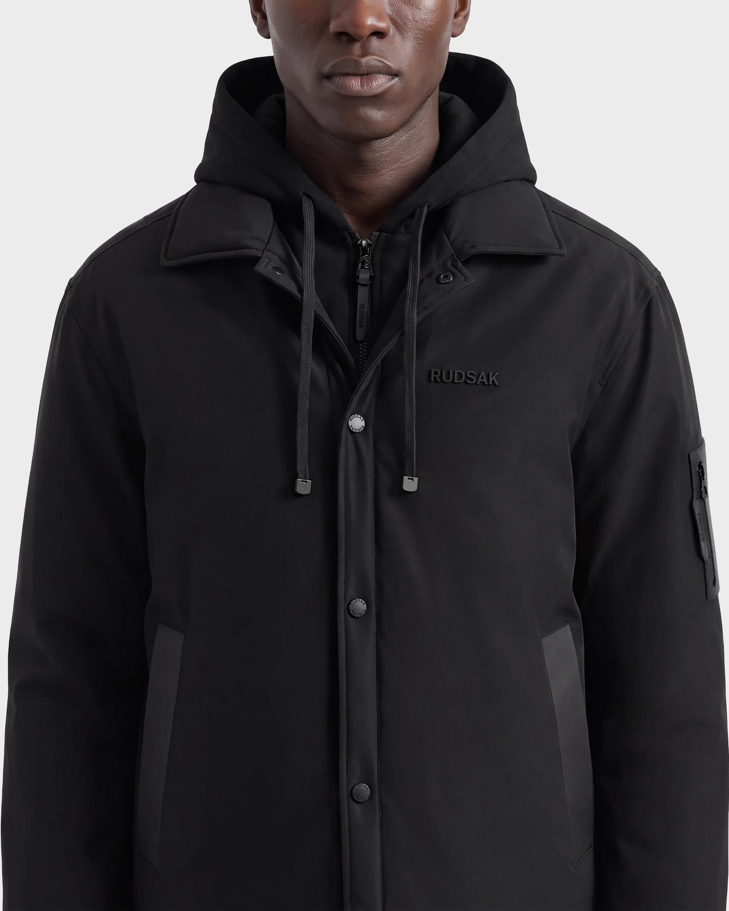 A person wearing the Rudsak LUCA - 6125009 Black men’s jacket, buttoned up, with a zipper pocket on the sleeve and drawstrings on the hood, is shown from the shoulders to mid-torso. This lightweight insulated outerwear combines stylish design with weather-resistant functionality.