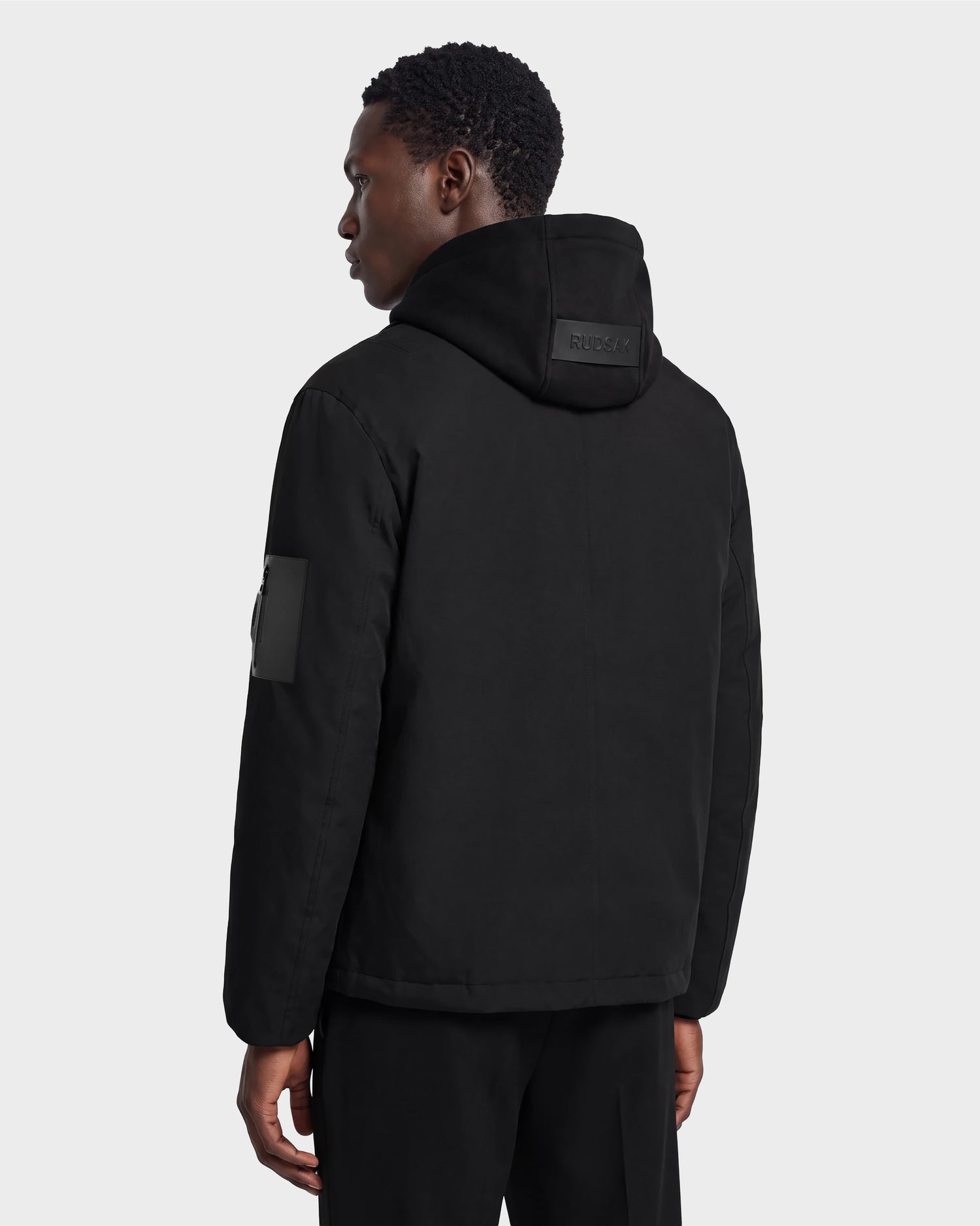 A person stands facing away from the camera, wearing the Rudsak LUCA - 6125009 Black weather-resistant men’s jacket and black pants against a plain background.