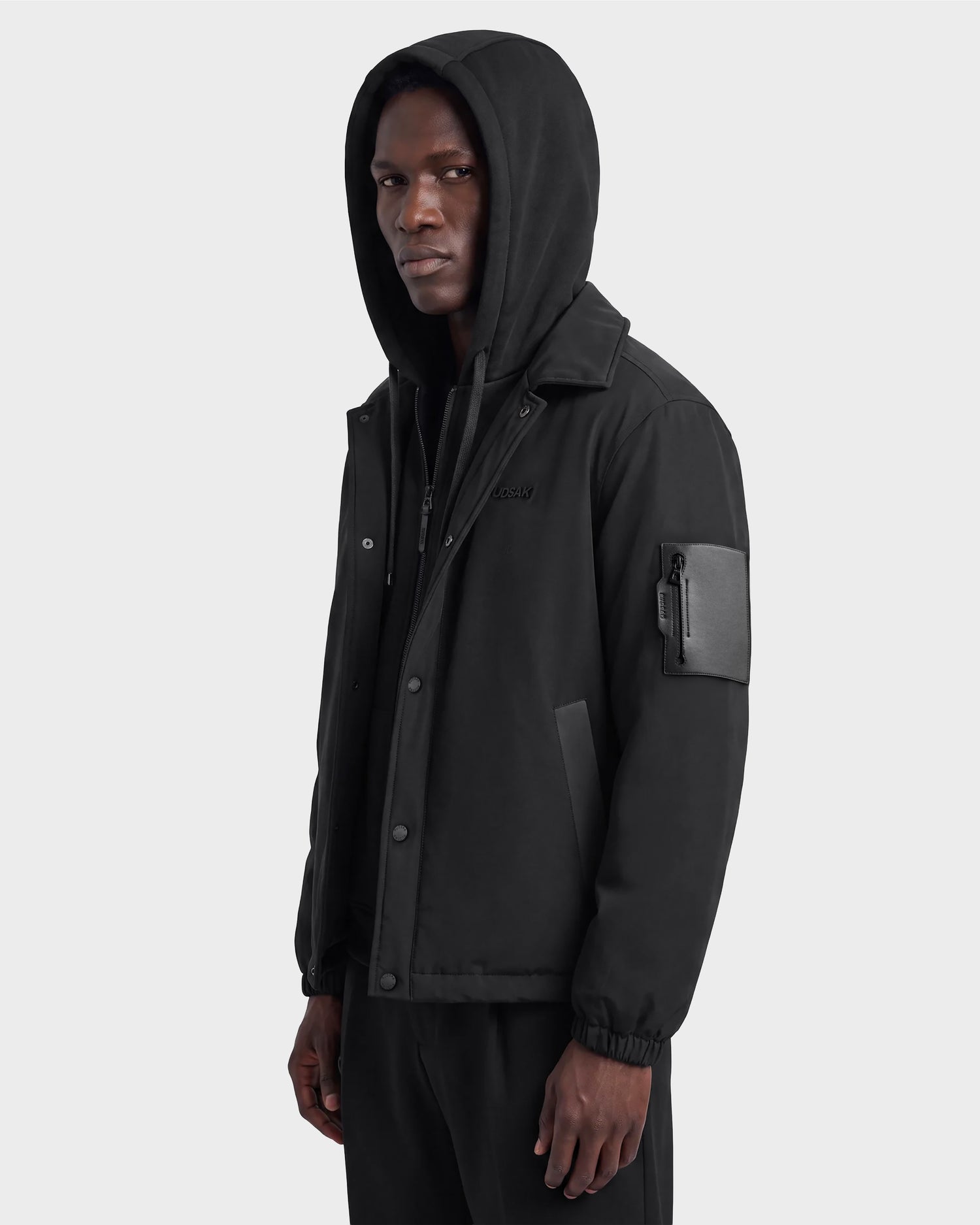 A person wearing the Rudsak LUCA - 6125009 Black, a weather-resistant hooded jacket featuring a zippered sleeve pocket, stands against a plain background, looking slightly to the side.