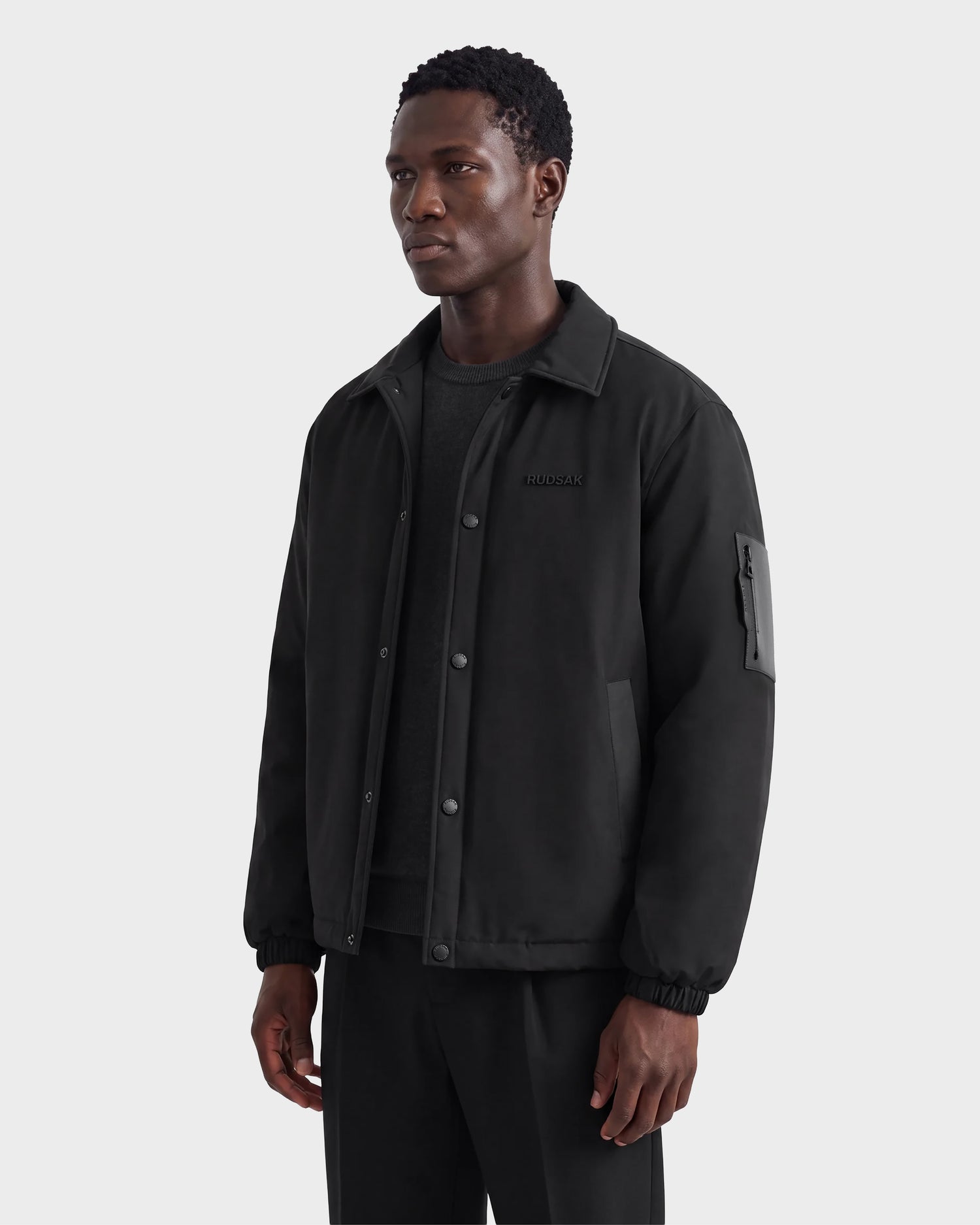 A man wearing the Rudsak LUCA - 6125009 Black weather-resistant men’s jacket over a dark shirt and pants stands against a light gray background, looking slightly to the side with a neutral expression.