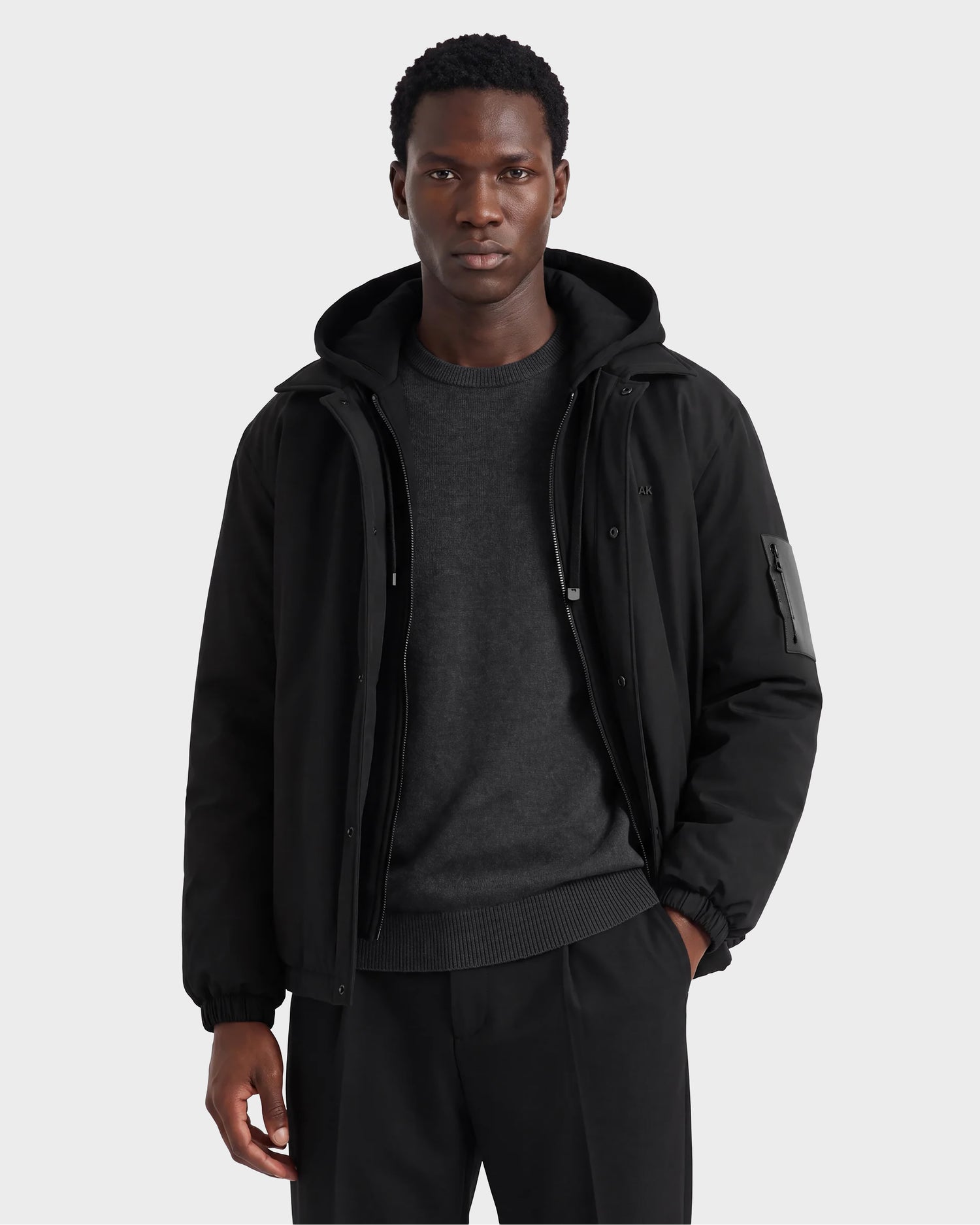 A man stands against a light gray background wearing the Rudsak LUCA - 6125009 Black weather-resistant men’s jacket over a dark gray sweater and black pants, with his left hand in his pocket, looking directly at the camera.
