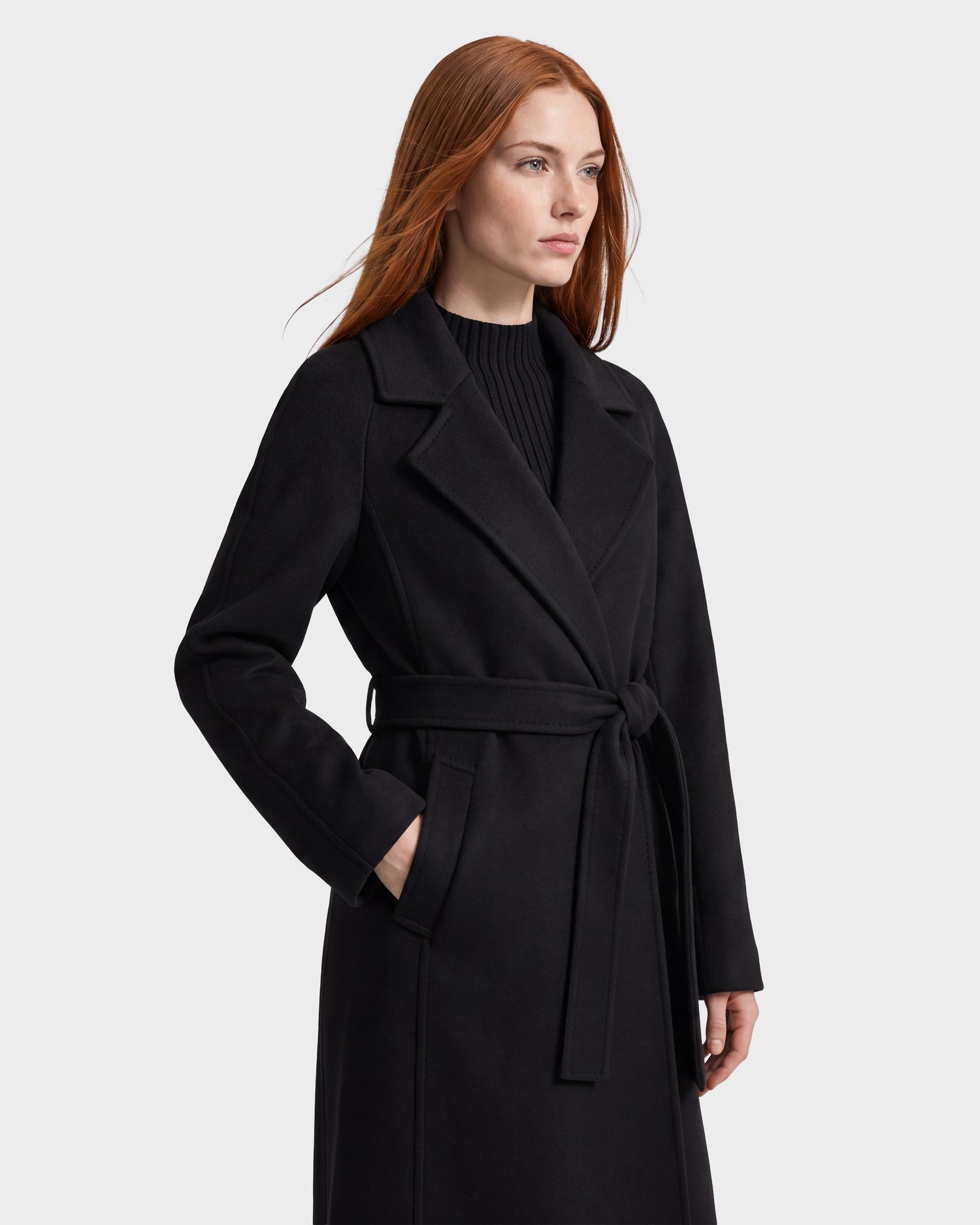 A woman with long red hair stands against a plain light gray background, looking to the side with one hand in her pocket, wearing the Rudsak LILIA - 8125634 black wool silk cashmere coat.