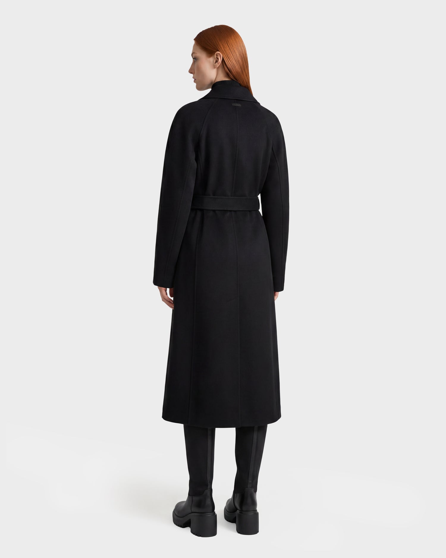 A person with long red hair stands facing away against a plain light gray background, wearing the Rudsak LILIA - 8125634 Black women’s wrap coat, belted at the waist, paired with black boots.