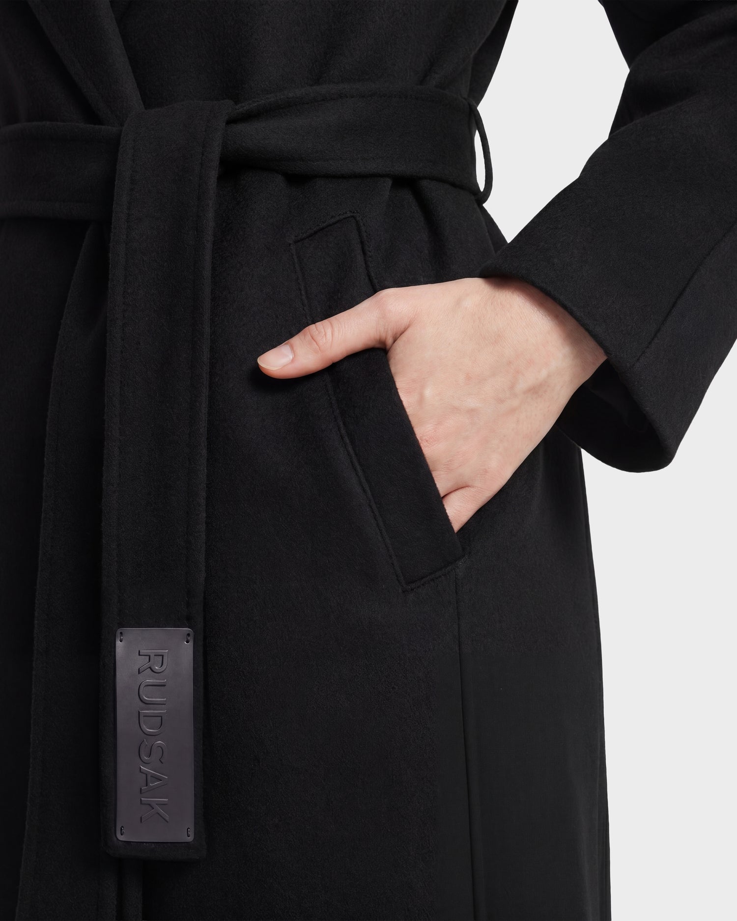 A person wearing the LILIA - 8125634 Black wrap coat by Rudsak, featuring a black tied belt with a leather tag embossed with the Rudsak logo, adds a refined touch to this elegant women's coat as they place their right hand in the pocket.