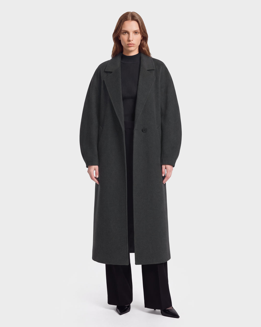A person stands facing forward, wearing the Rudsak LILIA - 8125634 Heather Charcoal long coat over a black ribbed sweater, black pants, and black pointed shoes, against a plain white background.