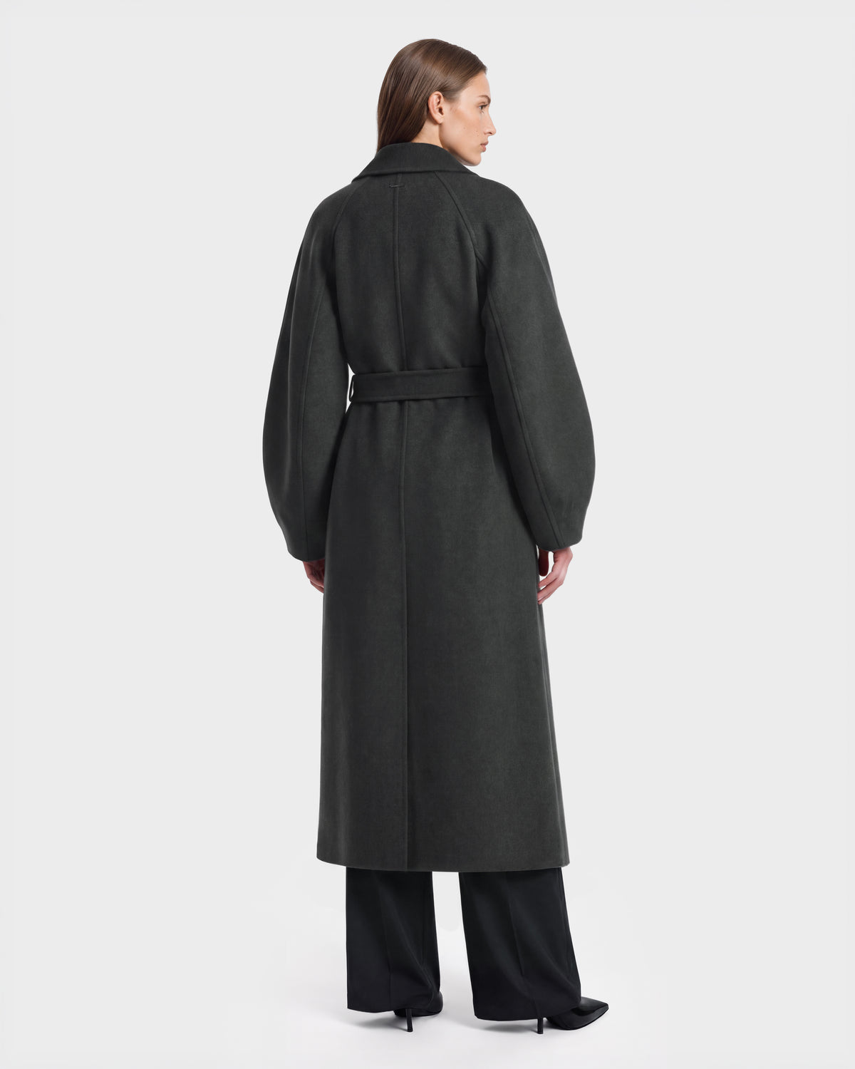 A woman stands facing away, wearing the Rudsak LILIA - 8125634 Heather Charcoal coat—a women’s long wrap style featuring a belted waist and wide sleeves—paired with black pants. The background is plain and light-colored.