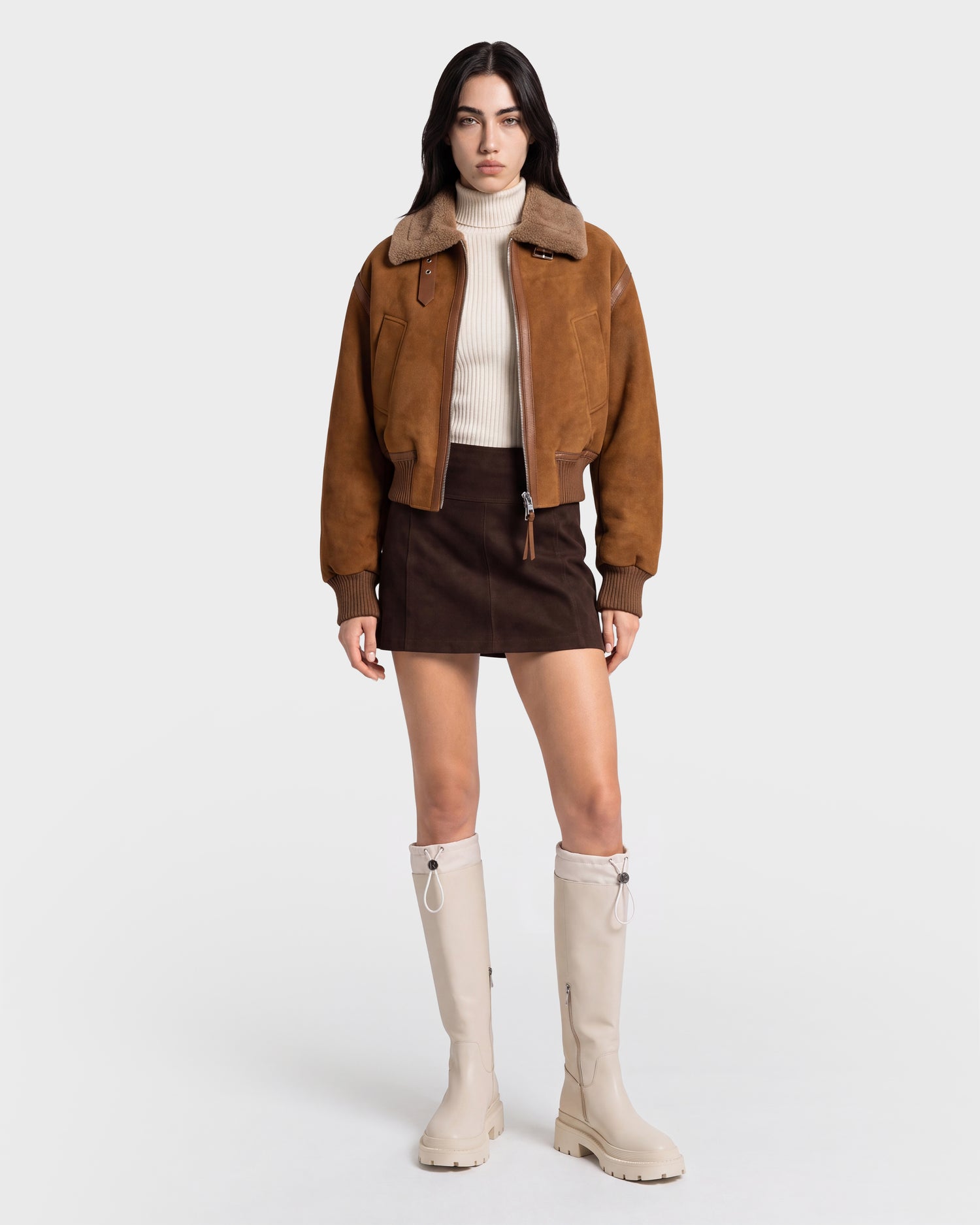 A woman stands against a plain background wearing the Rudsak LIZZIE - 8126787 Tan shearling jacket in a cropped fit. She pairs it with a cream turtleneck sweater, dark brown mini skirt, and knee-high beige boots, facing forward with a neutral expression.