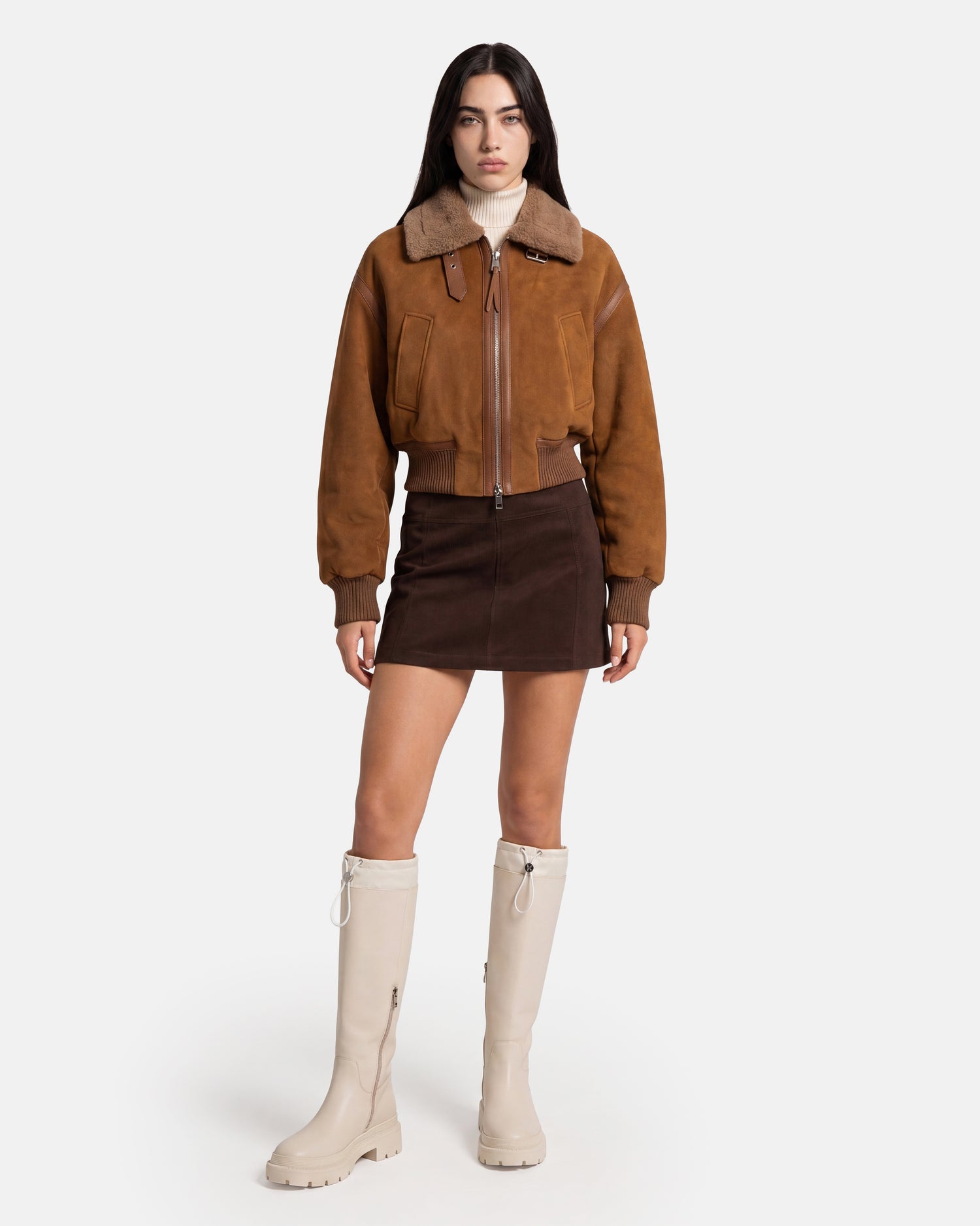 A woman stands against a plain white background wearing the Rudsak LIZZIE - 8126787 Tan cropped-fit brown shearling jacket, styled with a dark brown mini skirt, a light turtleneck, and knee-high beige boots crafted from soft suede.
