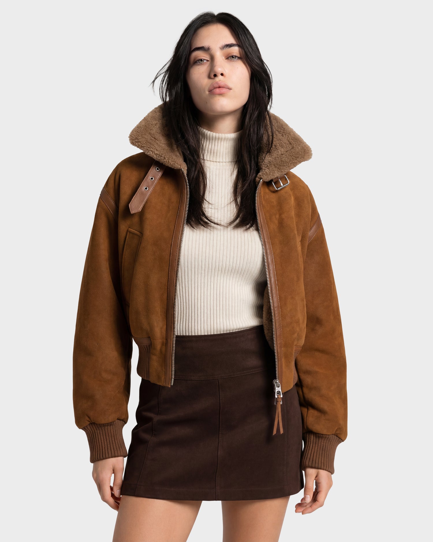 A woman stands confidently against a plain light background, wearing the Rudsak LIZZIE - 8126787 Tan soft suede shearling jacket in a cropped fit over a cream turtleneck sweater and a dark brown mini skirt, looking directly at the camera.