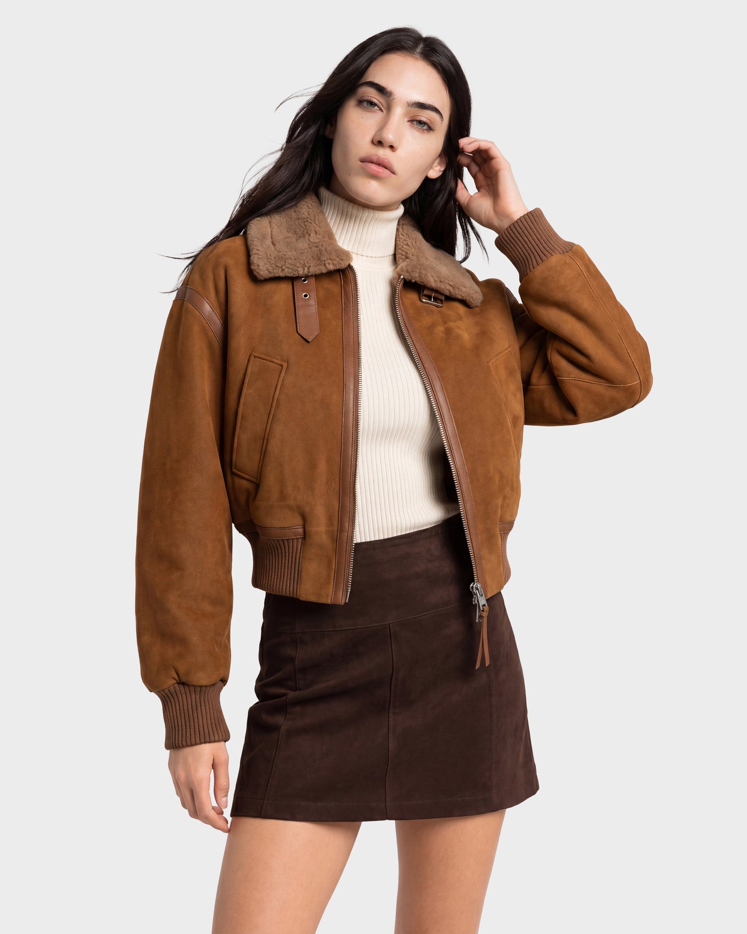 A woman stands against a light gray background wearing the LIZZIE - 8126787 Tan suede bomber jacket by Rudsak, featuring a shearling collar and cropped fit. She pairs it with a cream turtleneck sweater and a dark brown suede mini skirt.