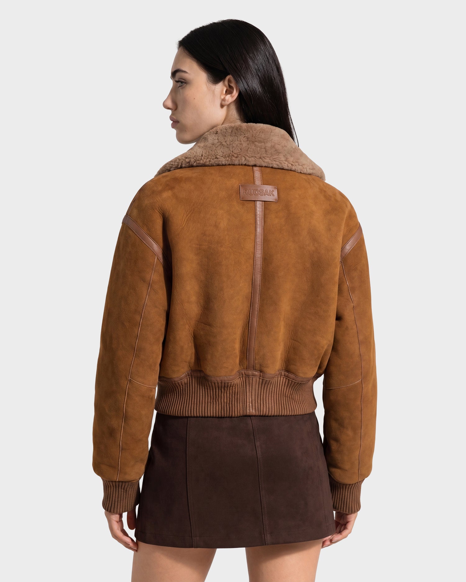 A woman with long dark hair wears the Rudsak LIZZIE - 8126787 Tan, a brown shearling jacket with a soft suede finish, paired with a dark brown skirt as she stands with her back to the camera against a plain light background.