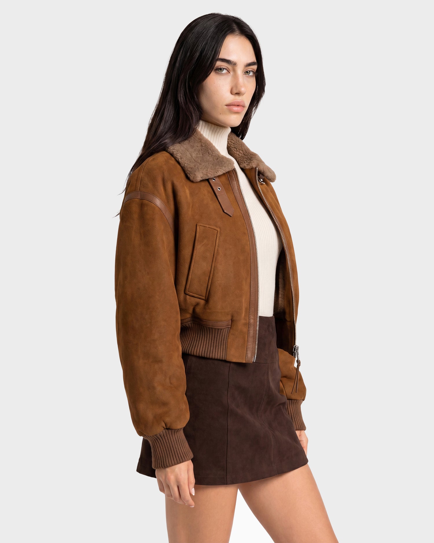 A woman stands against a plain light gray background wearing the Rudsak LIZZIE - 8126787 Tan suede bomber jacket, featuring a soft shearling collar and cropped fit, layered over a cream turtleneck and paired with a brown suede mini skirt.