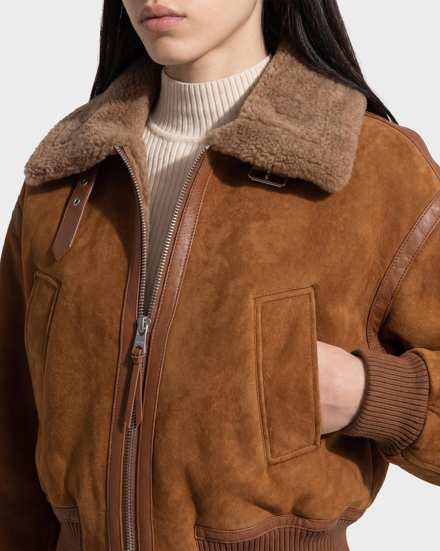 A woman wearing the Rudsak LIZZIE - 8126787 Tan shearling bomber jacket, featuring a brown suede finish with a soft tan wool collar and a cropped fit, styled over a beige ribbed turtleneck sweater, with her left hand tucked into the jacket pocket.
