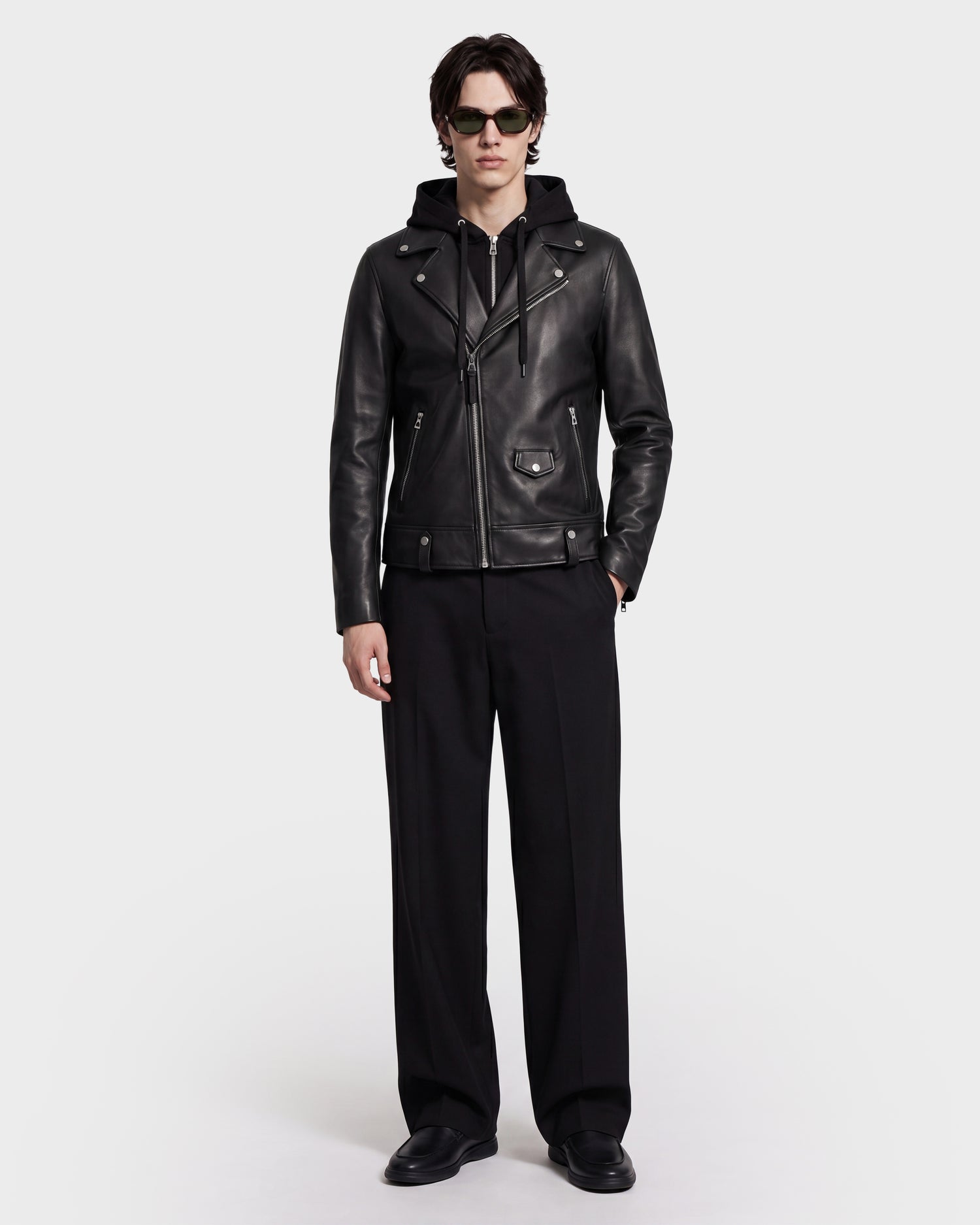 A person wearing sunglasses, black pants, and black shoes stands against a plain light background, facing forward with their hands by their sides, dressed in the Rudsak MANUS - 6124600 Black men's leather moto jacket featuring a removable hooded dickie.