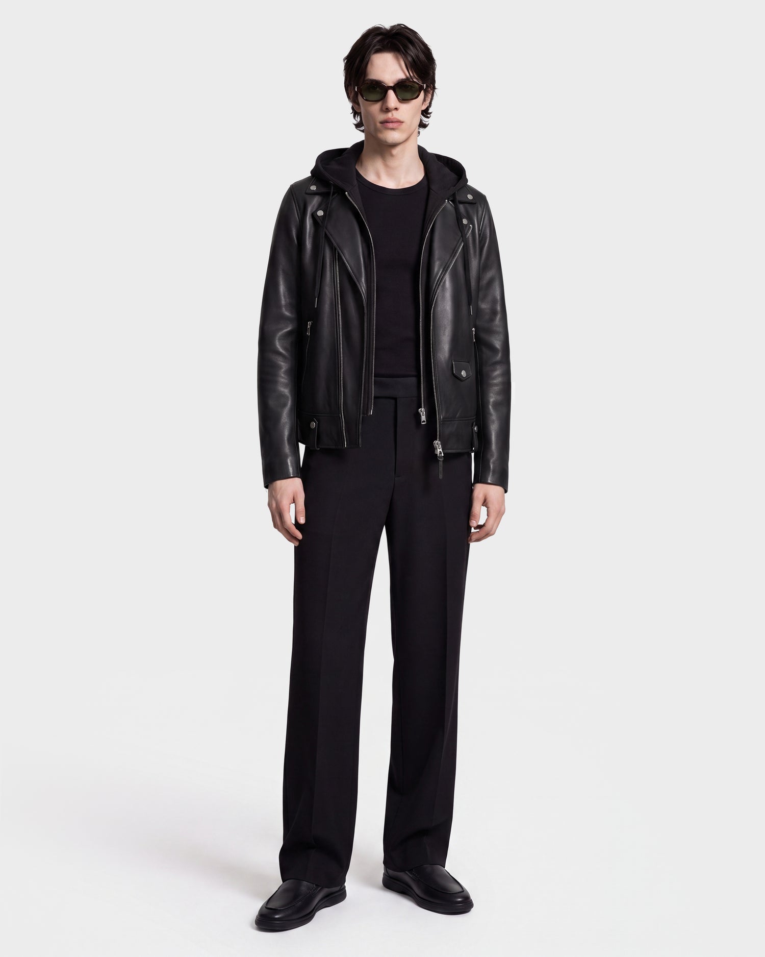 A person wearing sunglasses and the Rudsak MANUS - 6124600 Black men's leather moto jacket with a removable hooded dickie over a black shirt, paired with black pants and black shoes, stands against a plain white background.