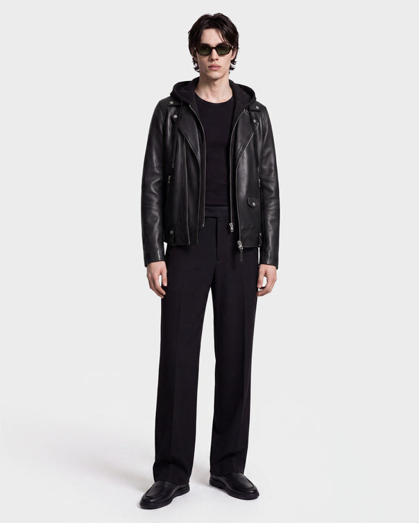 A person wearing sunglasses and the Rudsak MANUS - 6124600 Black men's leather moto jacket with a removable hooded dickie over a black shirt, paired with black pants and black shoes, stands against a plain white background.