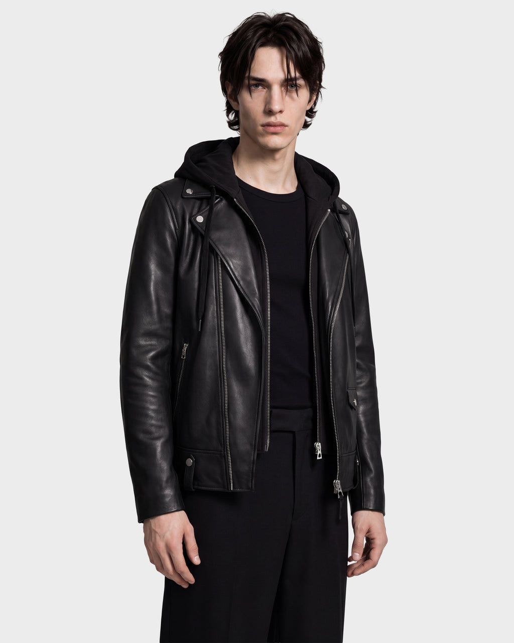 A person wearing the Rudsak MANUS - 6124600 Black men’s leather moto jacket with a removable hooded dickie over a black shirt and black pants stands facing forward against a plain light background.