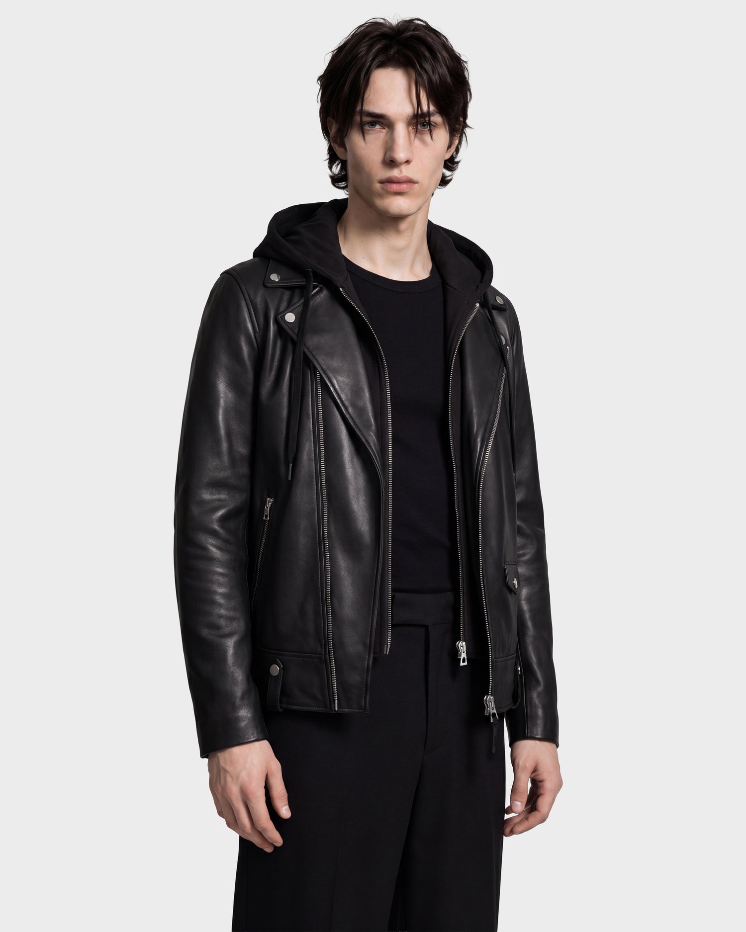 A young man with dark hair stands against a plain light background, looking directly at the camera with a neutral expression while wearing the Rudsak MANUS - 6124600 Black men’s leather moto jacket with a removable hooded dickie over a black hoodie and black pants.