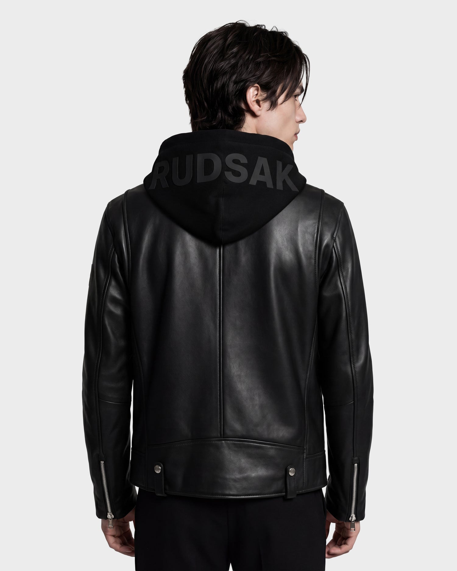 A person faces away, wearing the Rudsak MANUS - 6124600 Black men's leather moto jacket layered over a black hooded sweatshirt featuring "RUDSAK" in large letters on the hood. The plain light gray background highlights the jacket’s sleek biker style and its removable hooded dickie for added versatility.