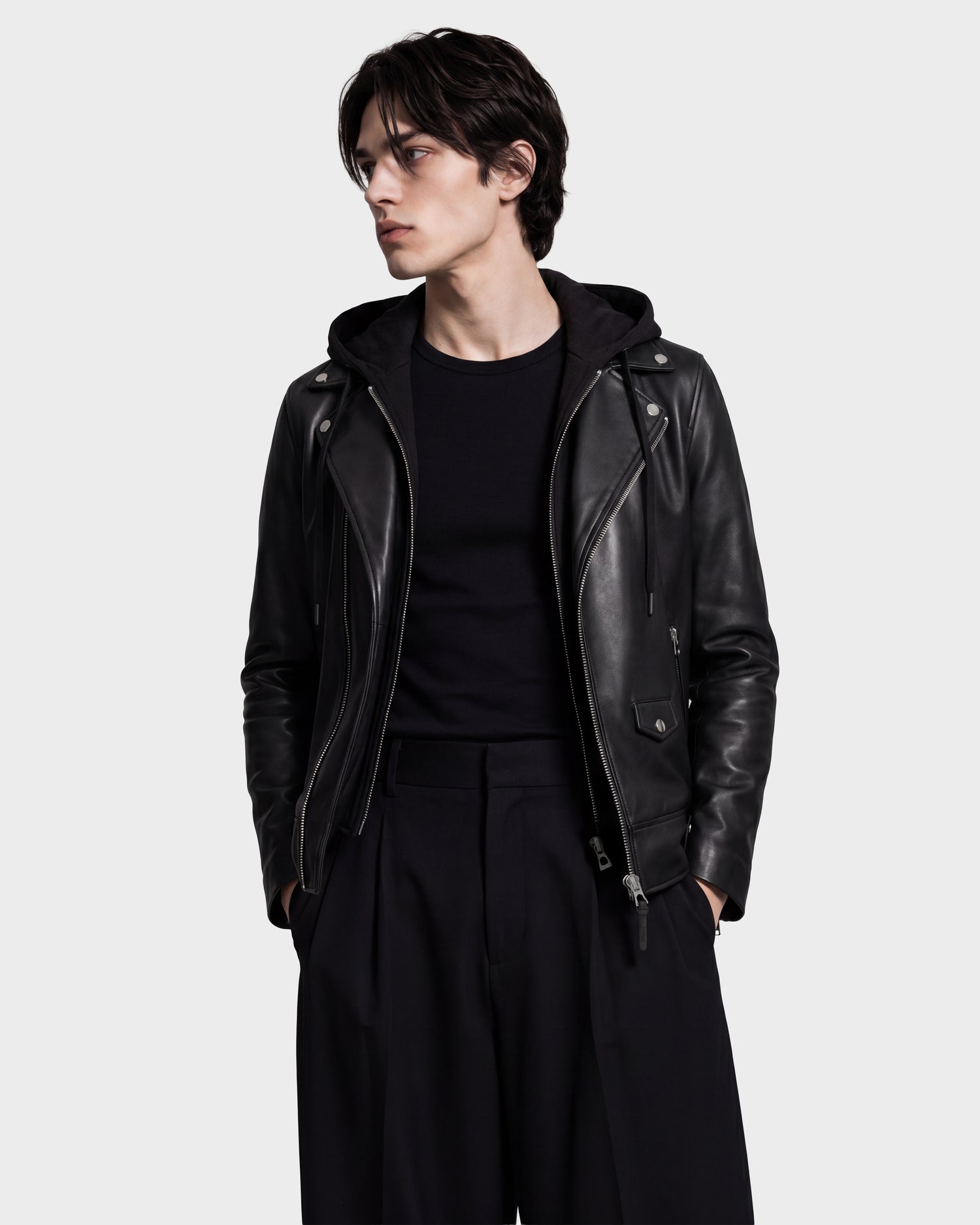 A person wearing the Rudsak MANUS - 6124600 Black men's leather moto jacket over a black shirt and black pants stands with hands in pockets, looking to the side against a plain background.