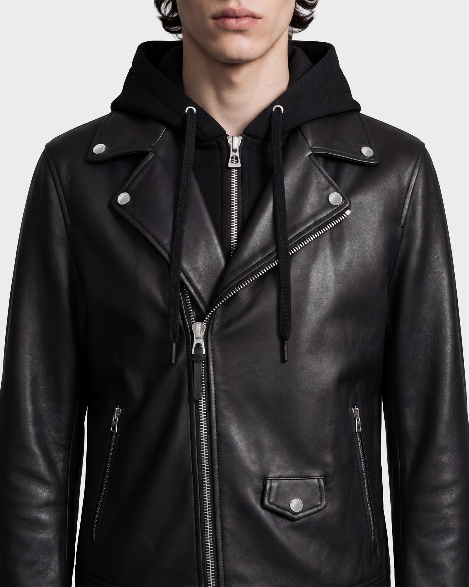 A person wearing the Rudsak MANUS - 6124600 Black men's leather moto jacket over a black hooded sweatshirt with silver zippers and buttons, shown from shoulders to mid-torso. This versatile biker jacket features a removable hooded dickie for added style and functionality.