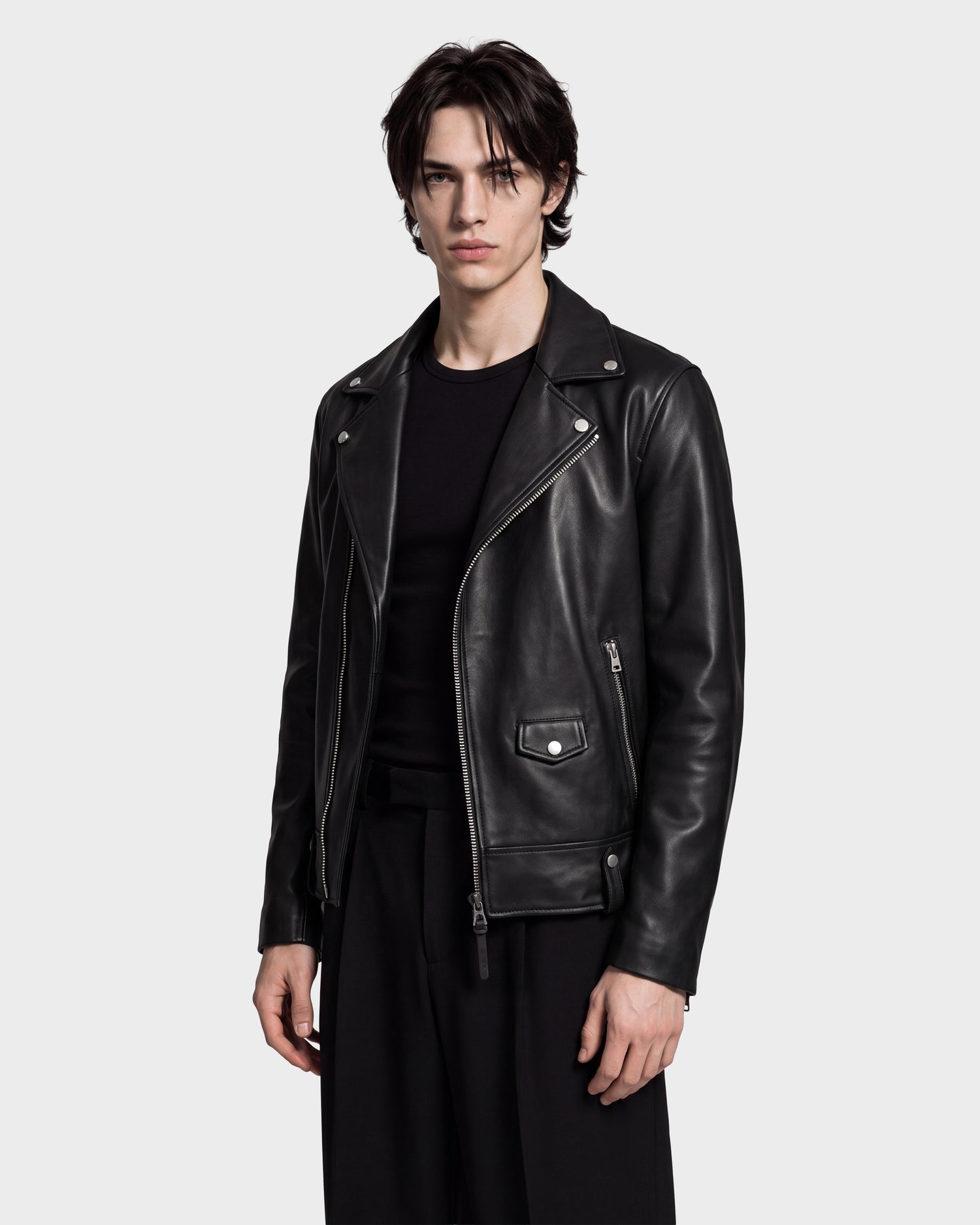 A person with dark hair wears the Rudsak MANUS - 6124600 Black men's leather moto jacket, which features a removable hooded dickie, over a black shirt and black pants while standing against a plain light gray background.