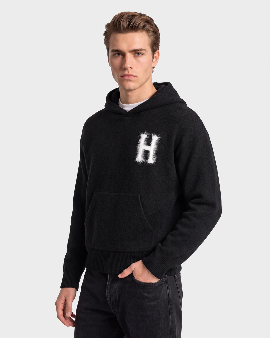 A person wearing the Rudsak MARCUS HV - H525503 Black hoodie, featuring a white “H” design on the chest, stands against a plain light background and looks at the camera with a neutral expression, effortlessly showcasing collegiate style.