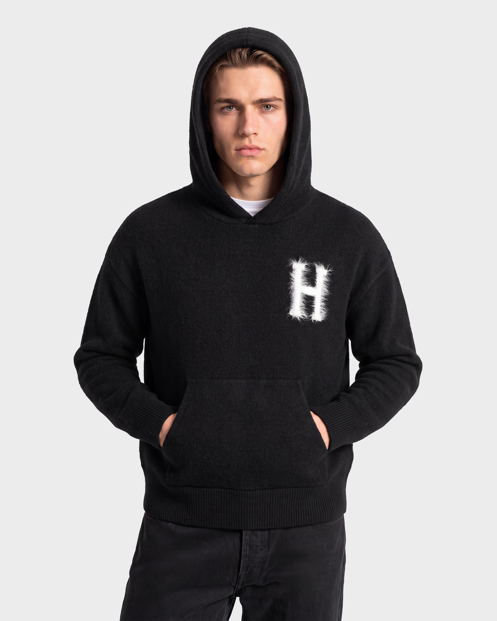 A person wearing the Rudsak MARCUS HV - H525503 Black hoodie, featuring a prominent H on the chest, stands with hands in the front pocket facing forward against a plain light gray background, showcasing a timeless collegiate style.