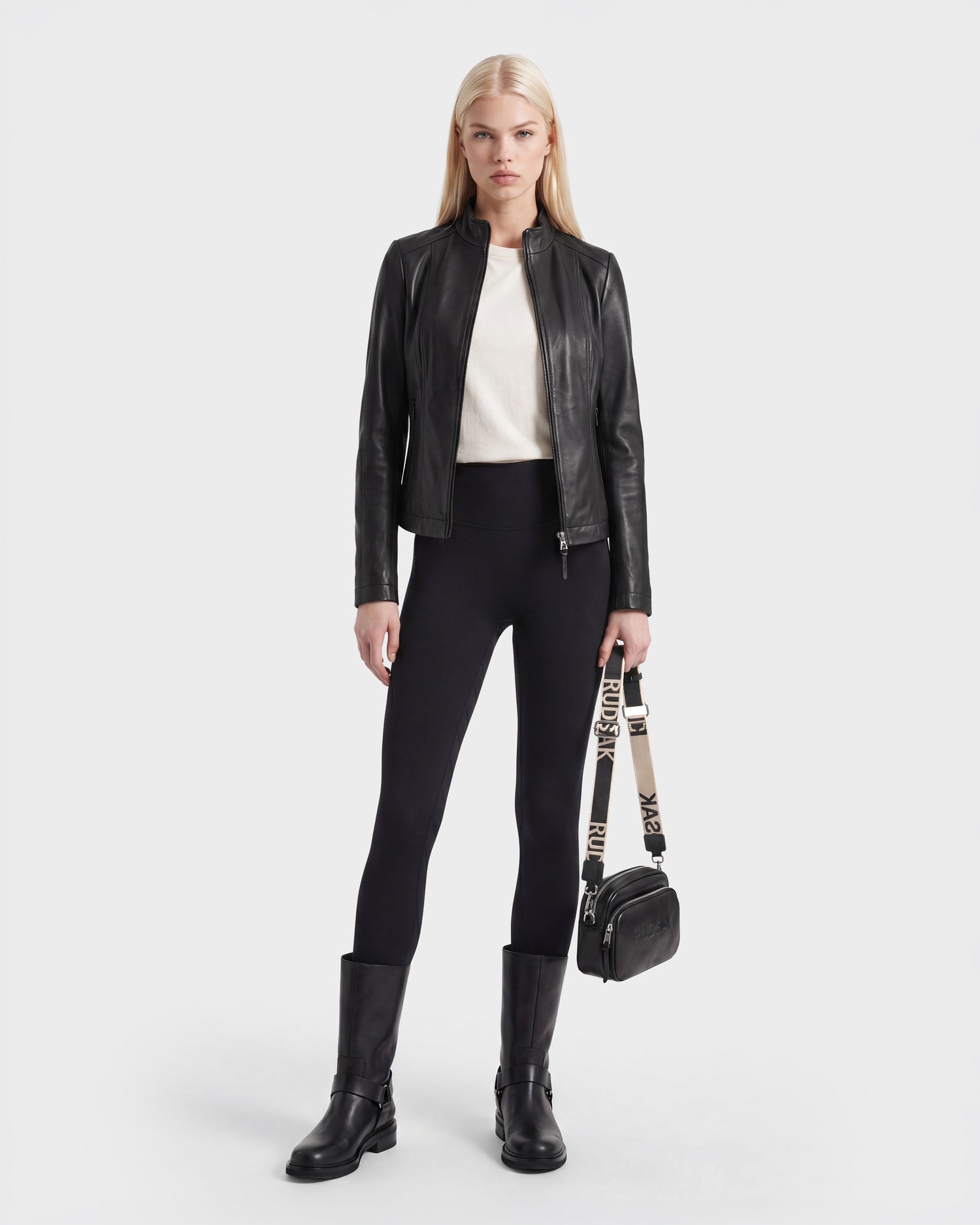 A person stands facing forward, wearing the Rudsak MARIAH - L124511 Black fitted moto biker jacket in black leather over a white top, paired with black leggings and black boots, while holding a small black handbag with a beige strap. The background is plain white.
