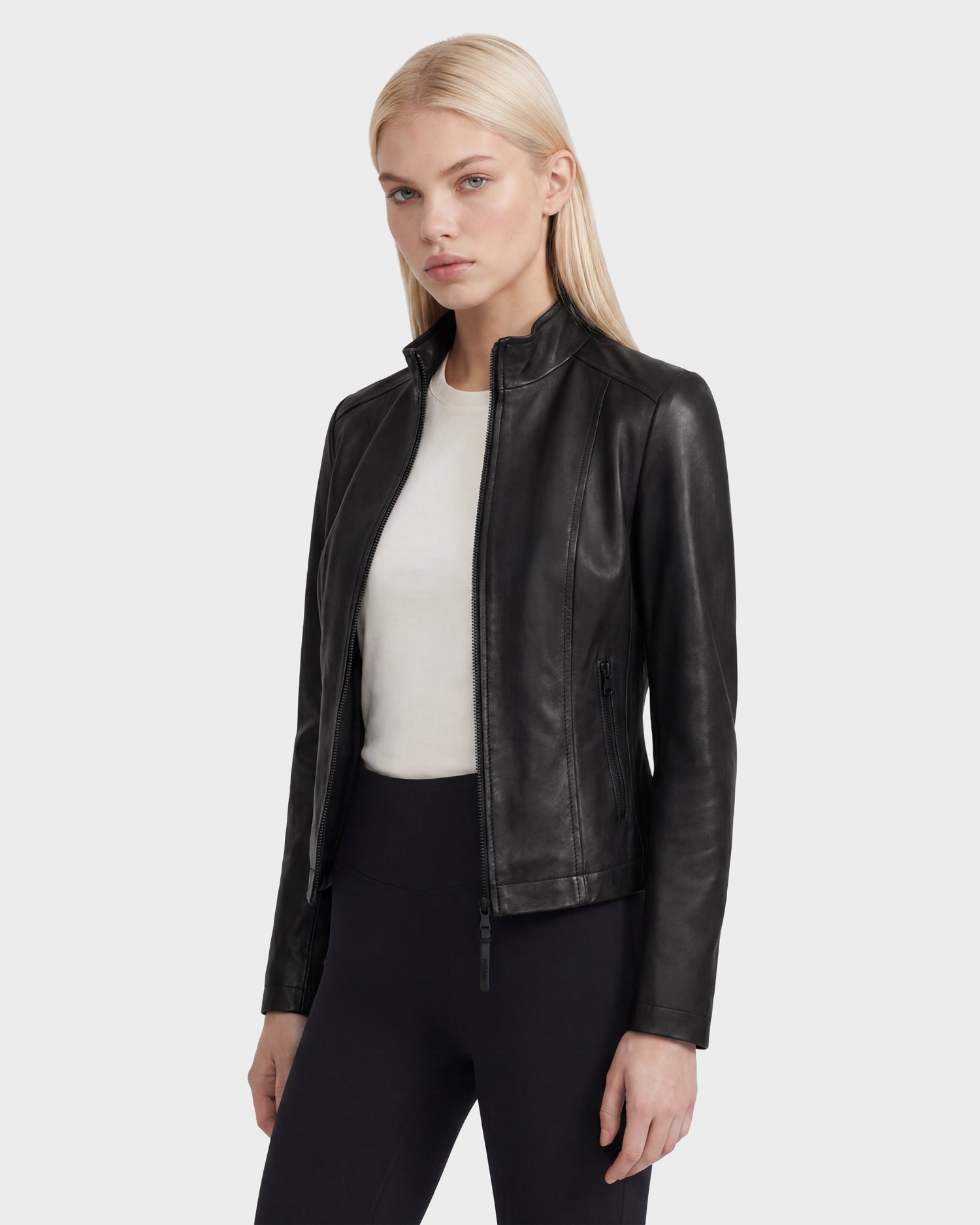 A woman with long blonde hair wearing the Rudsak MARIAH - L124511 Black fitted women’s leather jacket over a white top and black pants, standing against a plain light background.